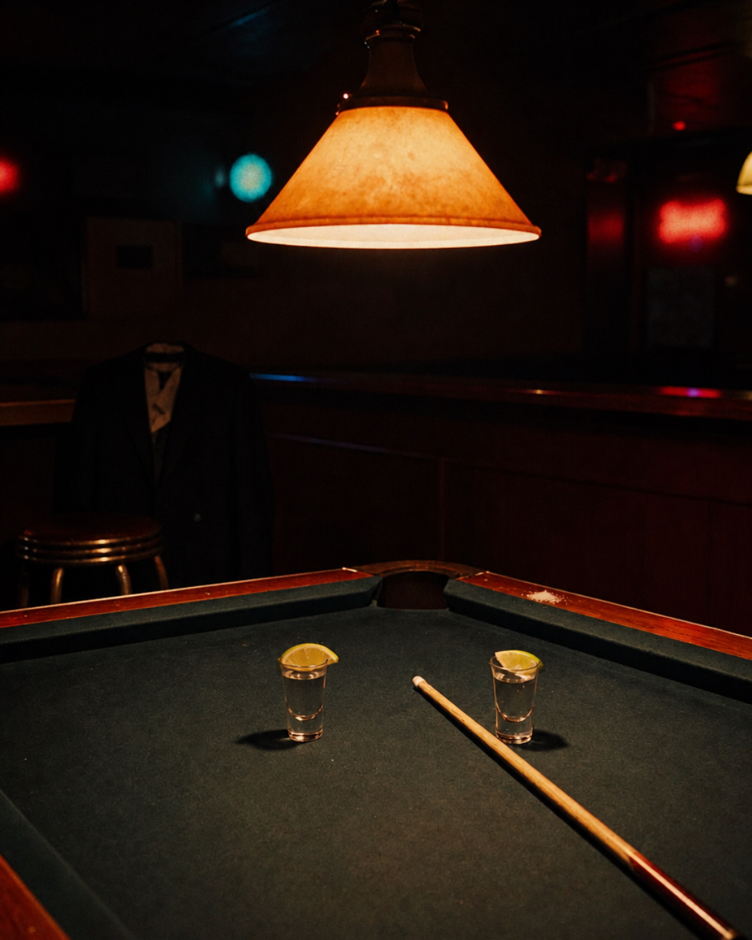Two tequila shots and a pool cue on a pool table in a dim bar at night