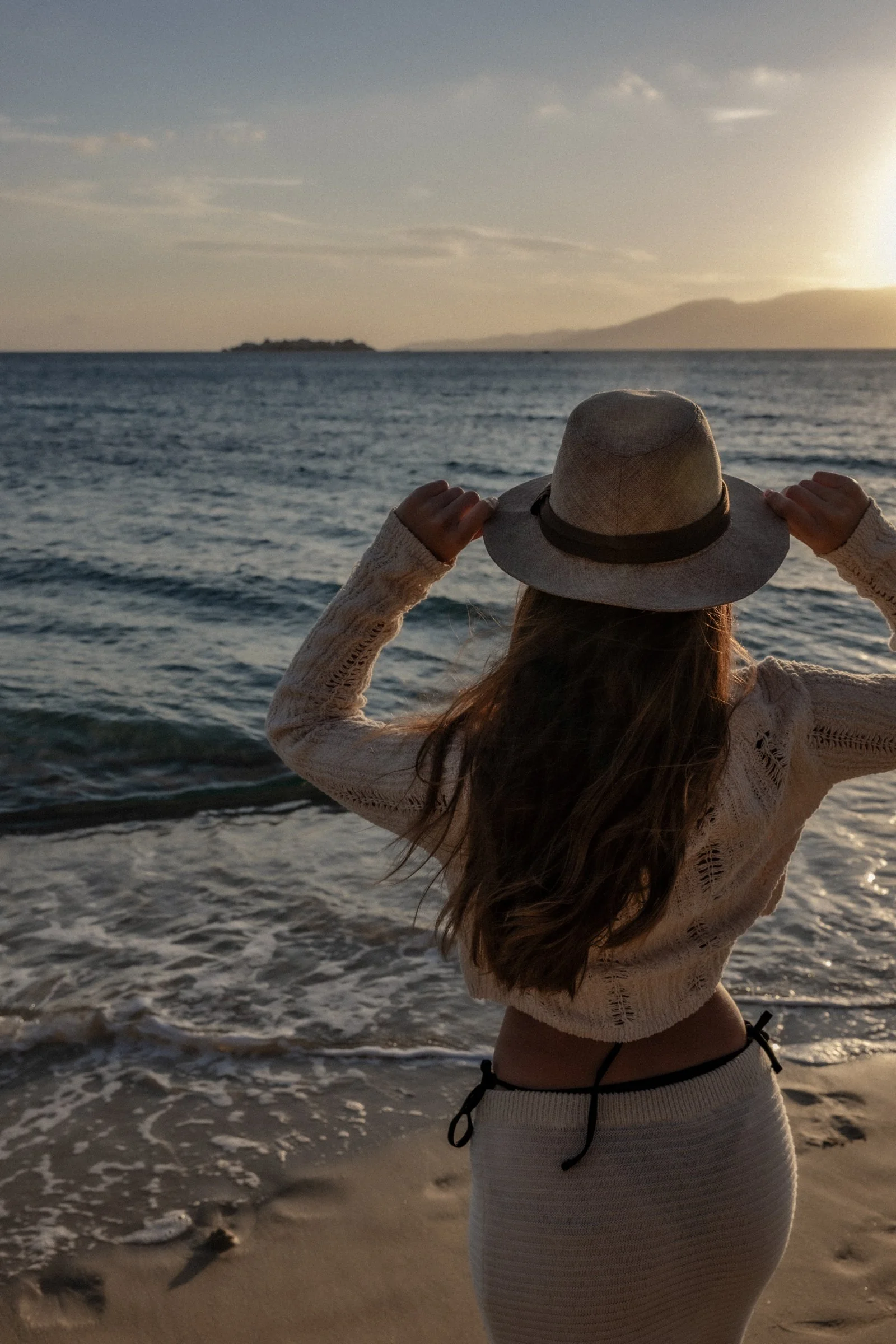 A woman standing on the beach at sunset, facing the ocean, holding a wide-brimmed hat, with waves and rocks in the foreground and distant land and mountains in the background.