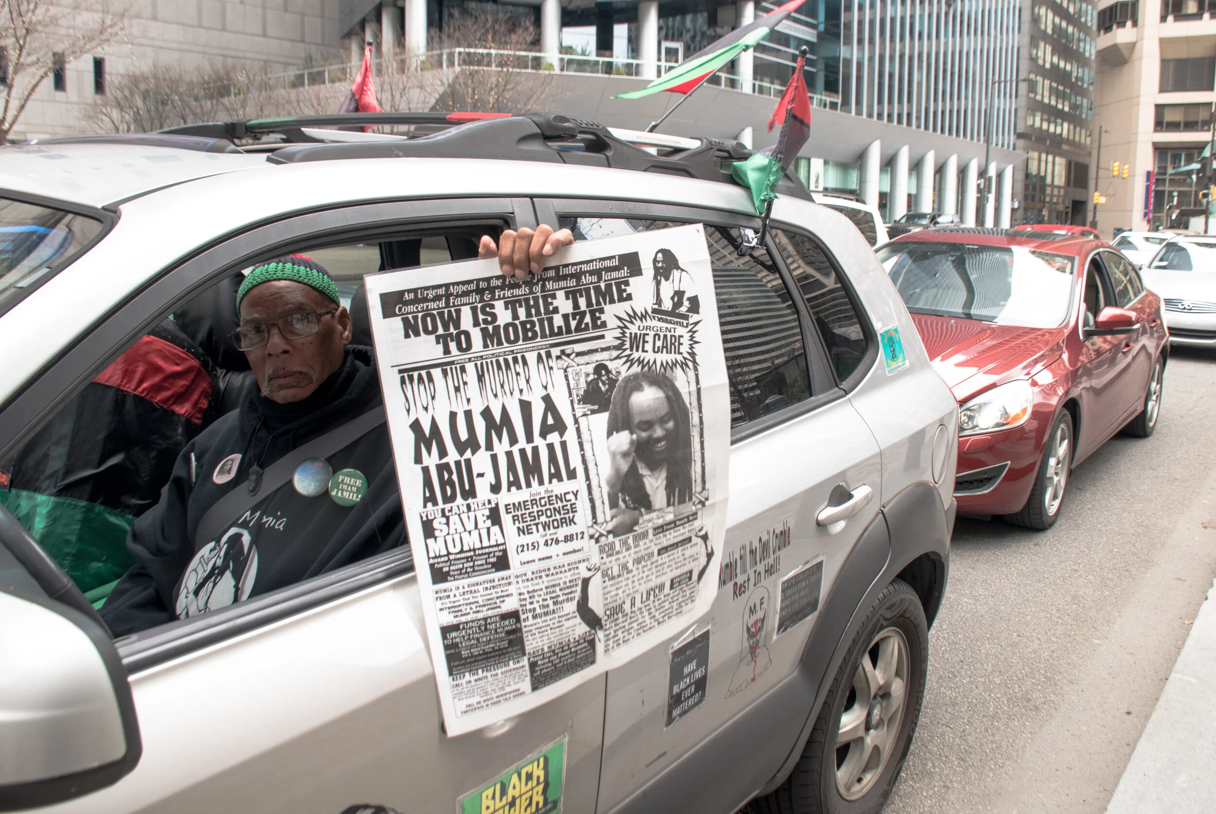 Mumia supporter Kamau Becktemba participating in an April 10 car caravan protest in Philadelphia. Traffic around Philadelphia City Hall came to a standstill for over an hour April 9 as hundreds of protesters from the safety of their cars demanded Mayor Jim Kenney and First Judicial District Judges Patrick Dugan, Leon Tucker, and Idee Fox decrease the numbers of incarcerated people in the city's jails.  Just days after being bailed out by the Philadelphia Community Bail Fund, Luz Acevedo said over a sound system: “I don’t think we should die in jail like animals die in cages,” referring to the high risk of death prisoners face from the COVID19 pandemic.  Over 75 drivers then continued the protest by moving to the city's jail complex several miles away where a delegation of religious leaders held signs outside the stone and rusty bars barriers, giving speeches and saying prayers. Protesters applauded by sounding their horns, hoping the over 4,200 prisoners on the other side of the walls could hear them.  The #No215Jail Coalition is demanding the city release the vast majority of people by granting parole, lifting detainers, ending cash bail, reviewing everyone in detention for possible release, and prioritizing release for youth, elderly people, and people who are medically vulnerable.  More photos at https://www.flickr.com/photos/109799466@N06/albums/72157713838662682