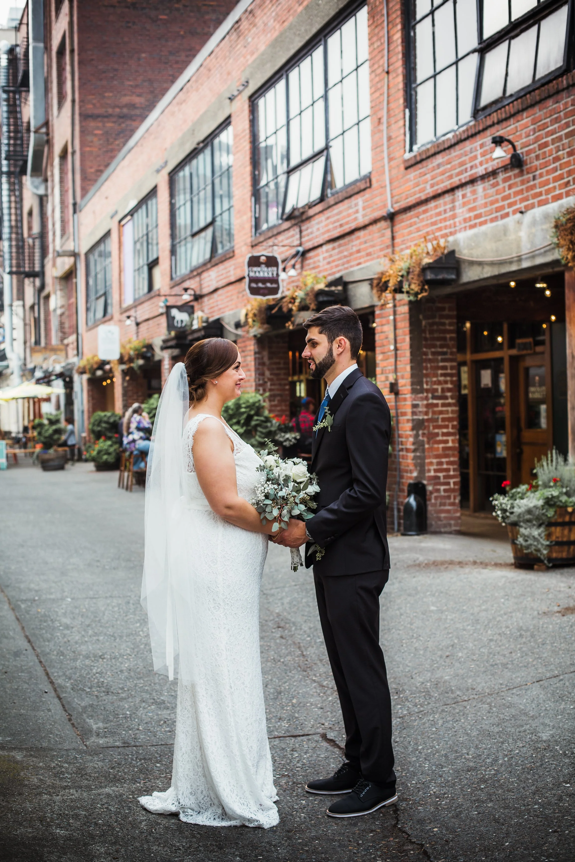 A bride and groom stand facing each other, holding hands, in a city street lined with brick buildings, during their wedding ceremony.