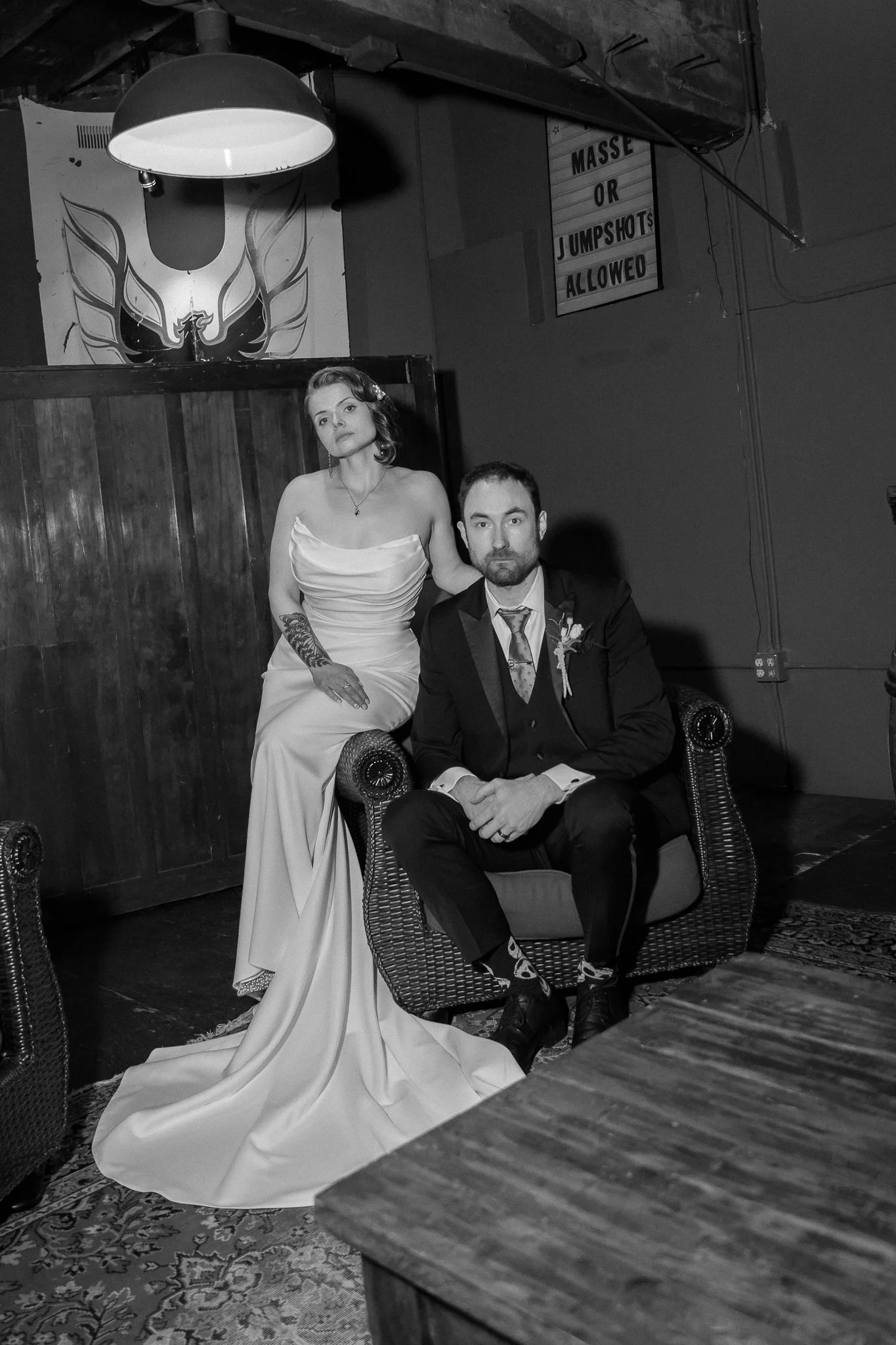 A woman in a wedding dress and a man in a tuxedo sitting on a wicker armchair in a dimly lit room, with a tattooed arm resting on her lap and a serious expression.