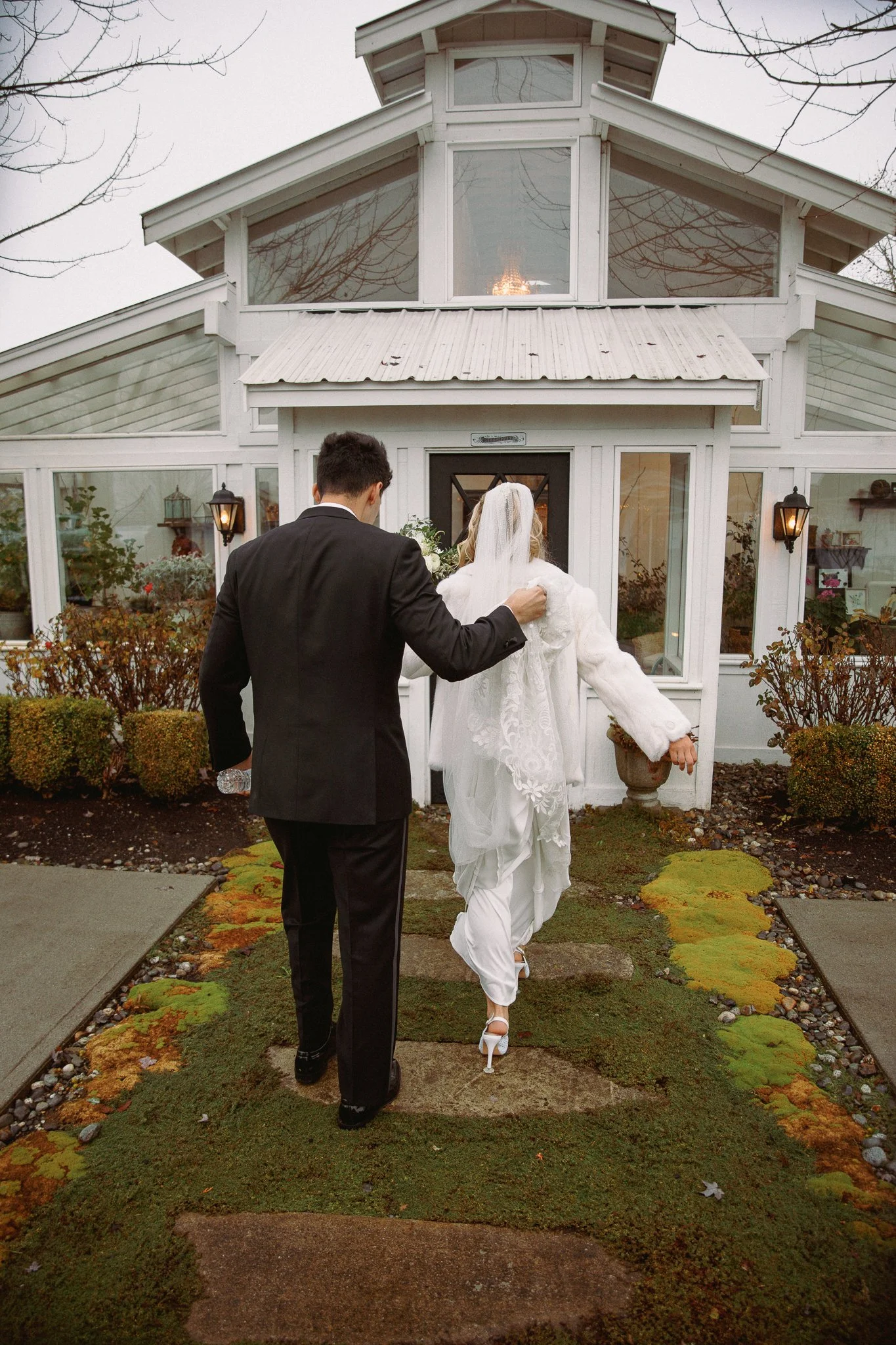 A bride and groom walking toward a white house or greenhouse exterior. The groom has his arm around the bride, who is wearing white high heels and a veil. The scene appears to be during late fall or winter, as the sky is overcast and the bushes outsi