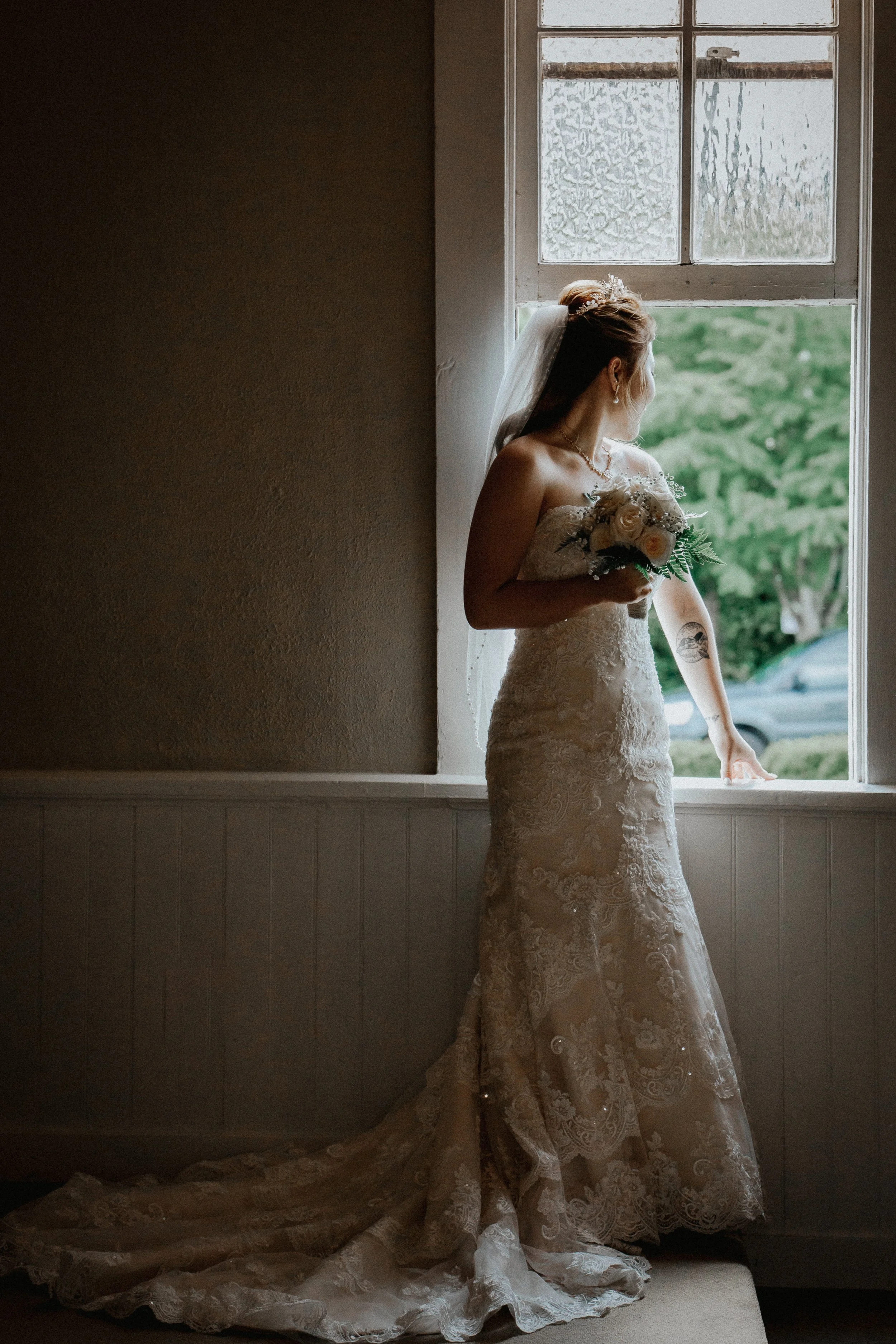 A bride in a lace wedding gown holding a bouquet of flowers, looking out a window with natural light illuminating her face.