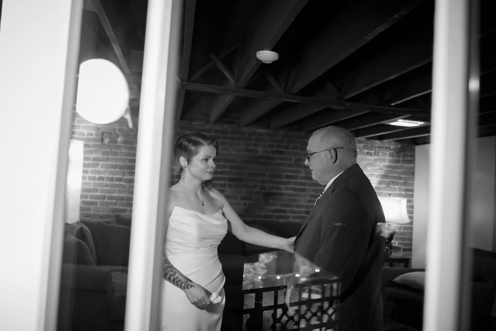 A black and white photo of a woman in a wedding dress holding a small cloth, looking at a man in a suit during a moment in a cozy room with brick walls, wooden ceiling beams, and a lamp in the background.