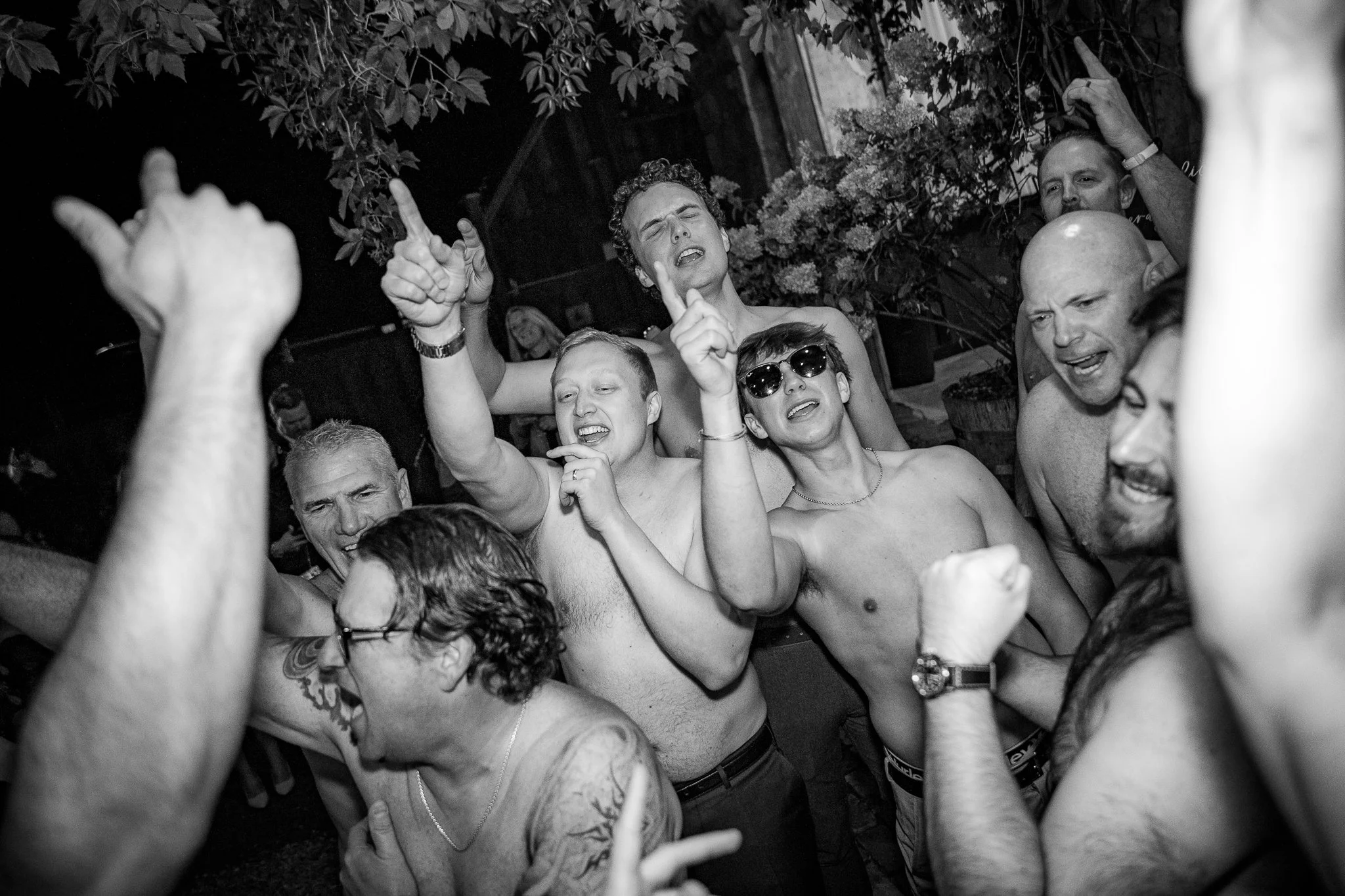 Group of shirtless men dancing and singing together at a lively party or celebration, some with sunglasses and tattoos, and everyone appears to be enjoying themselves.