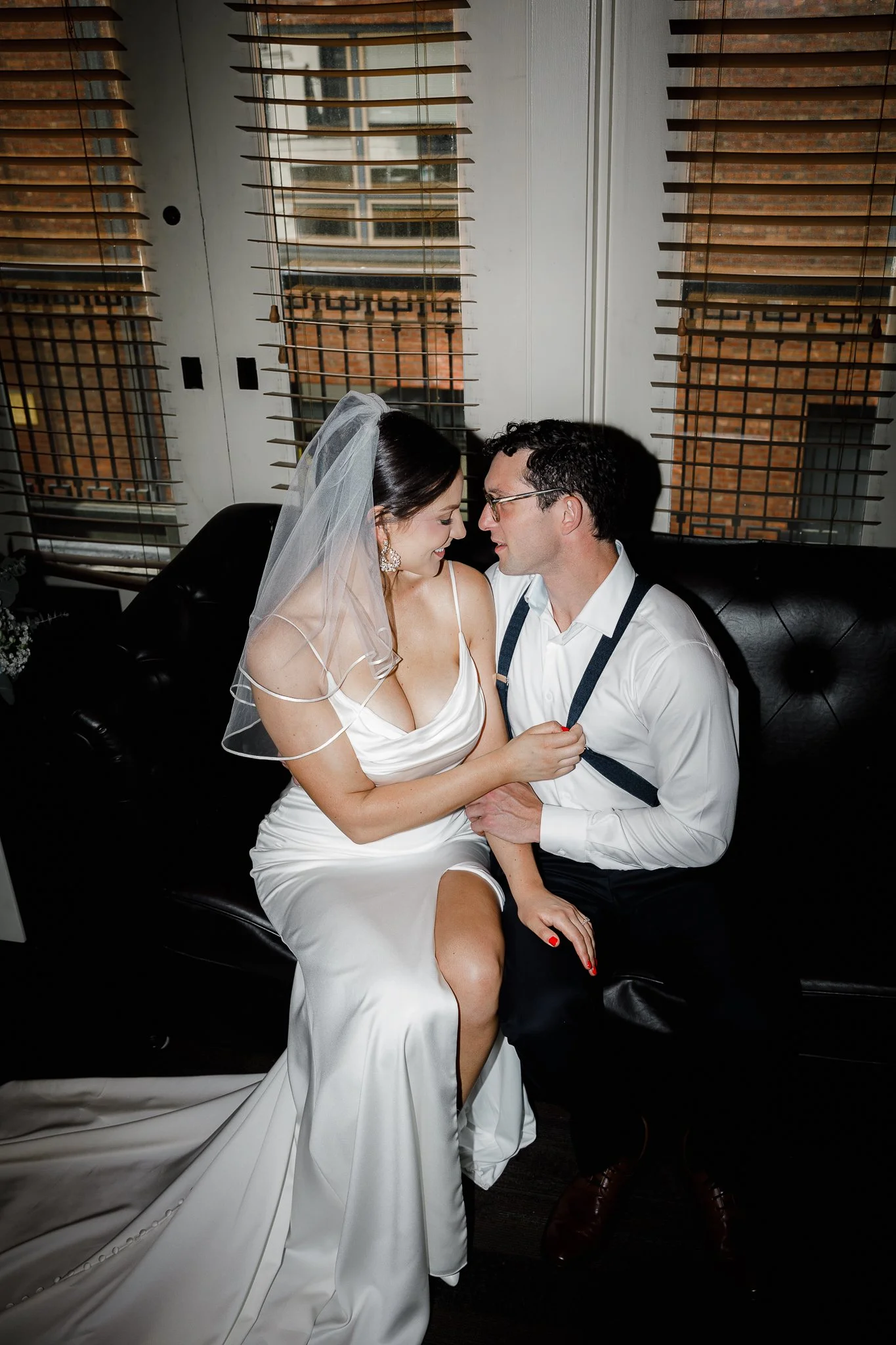A bride and groom sitting close together on a black leather couch, sharing an intimate moment indoors with wooden window shutters behind them.