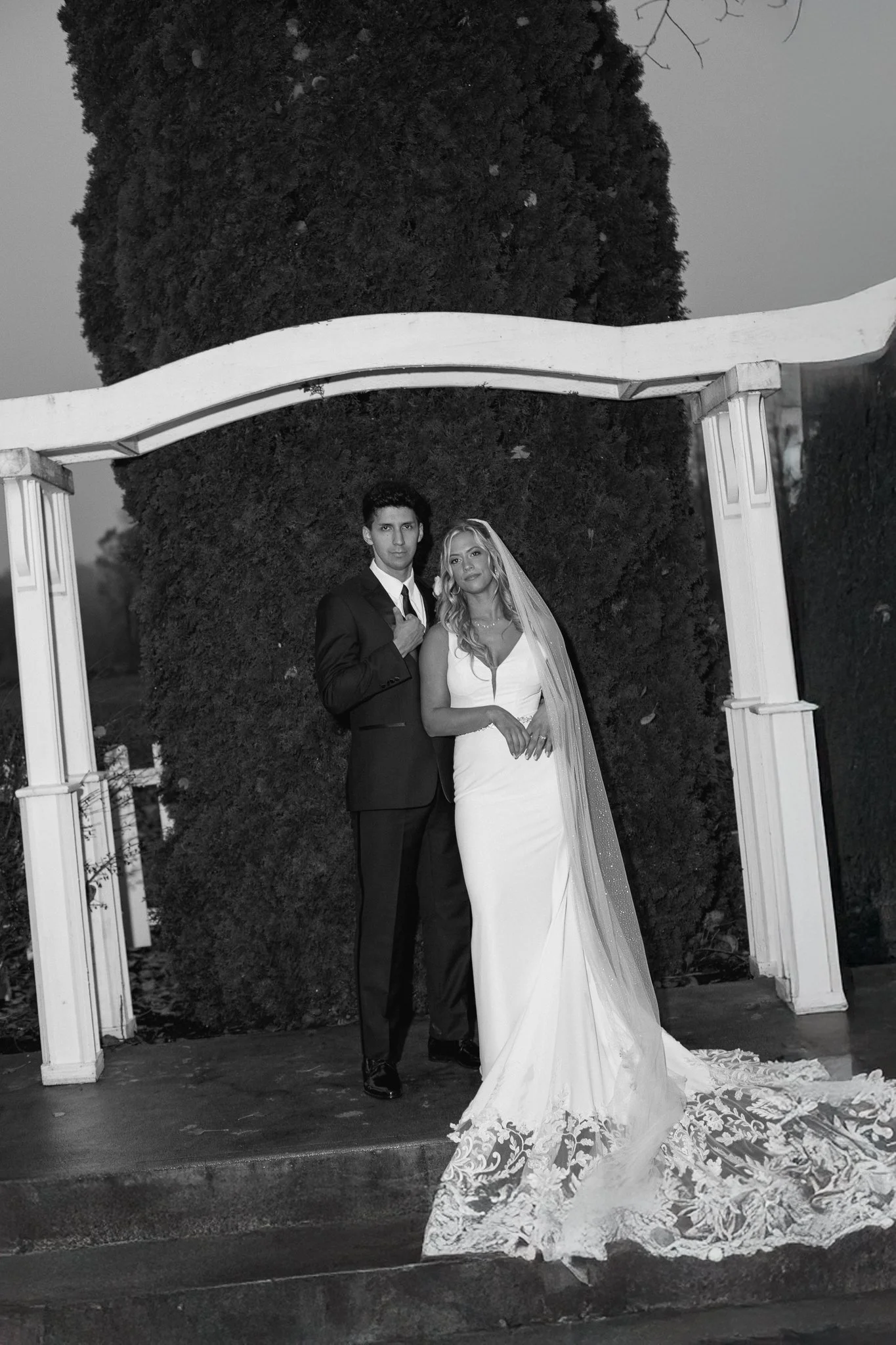 Black and white photo of a bride and groom standing together under a white wooden archway outdoors, with a large tree in the background. The bride is in a white wedding dress with lace details at the bottom, and the groom is in a dark suit with a tie