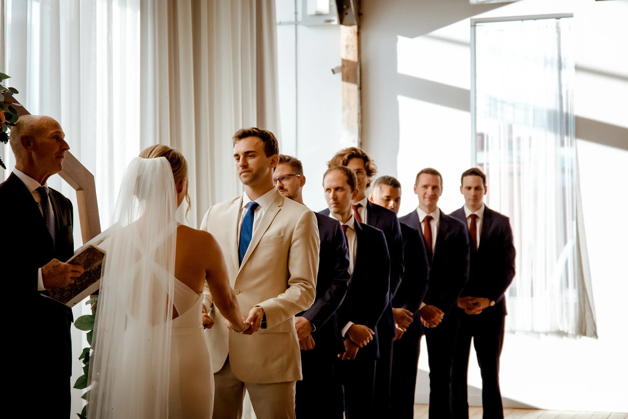 A bride and groom exchanging vows during their wedding ceremony, with a line of groomsmen standing behind them in a bright, modern venue.