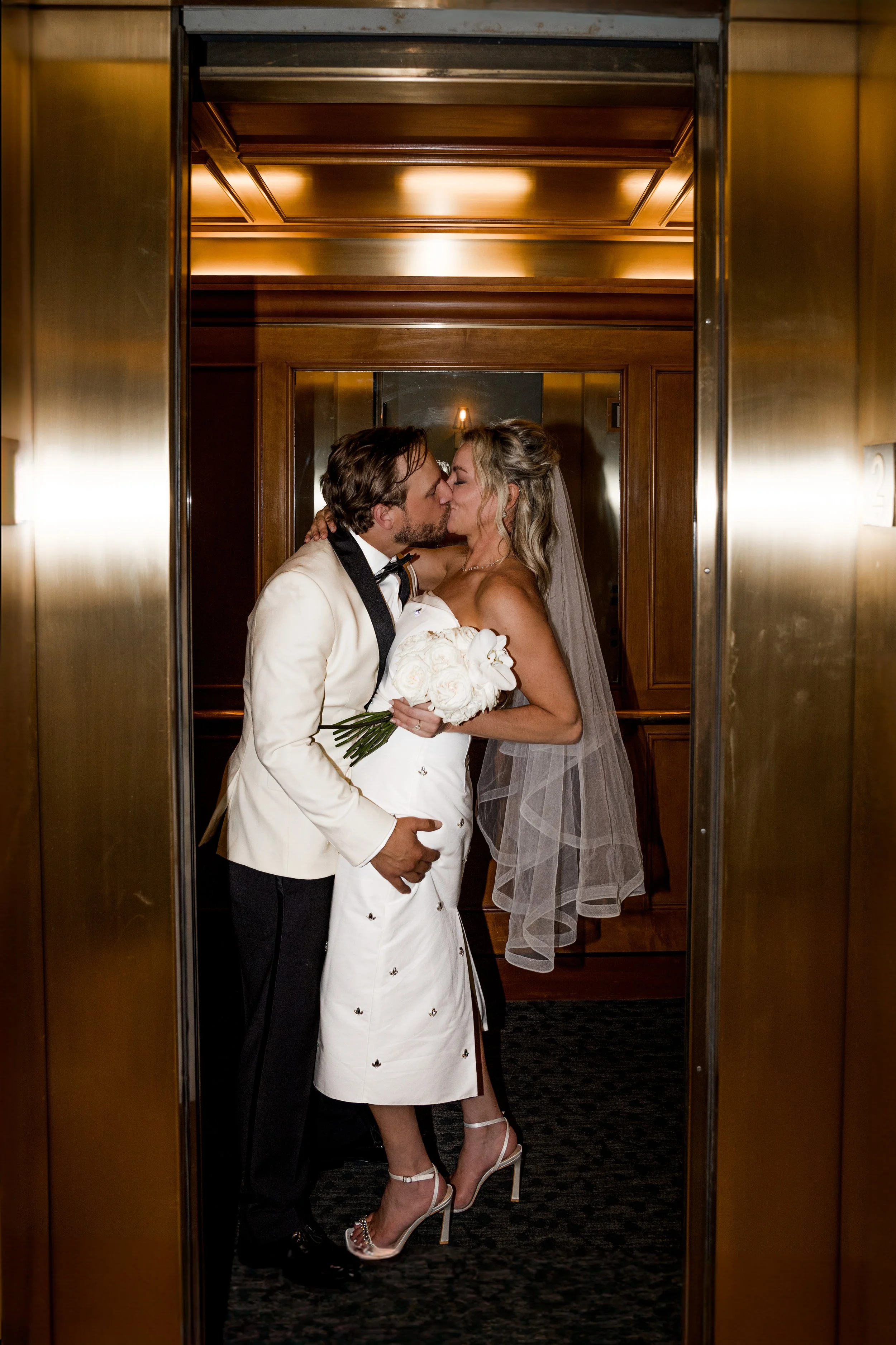 Bride and groom sharing a kiss in an elevator at their wedding, with the groom holding a bouquet of white roses.