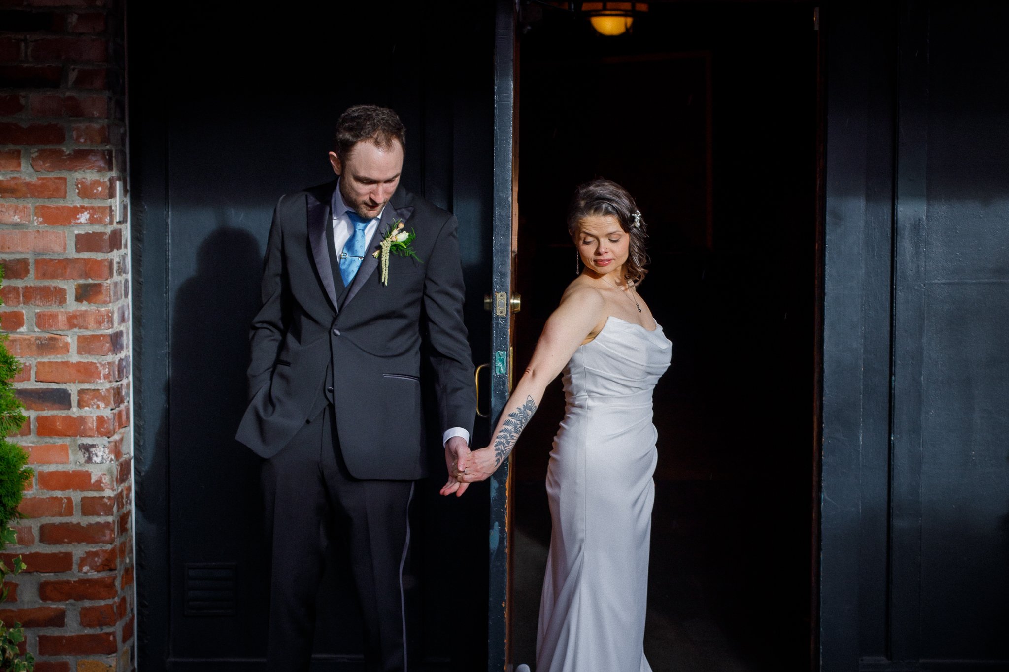 A bride and groom holding hands at their wedding, standing apart but connected, in front of a dark background with a brick wall on the side.
