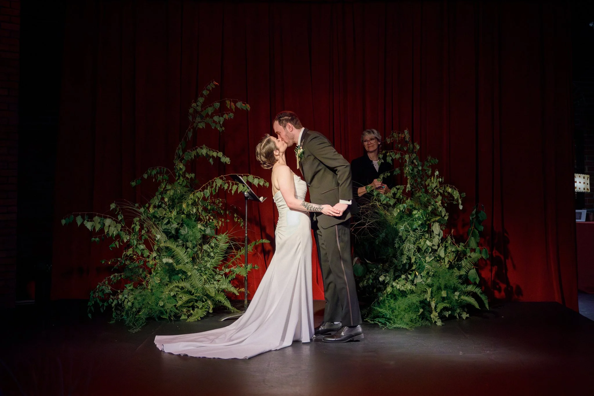 A bride and groom share a kiss during their wedding ceremony on a stage with red curtains and lush green plants in the background.