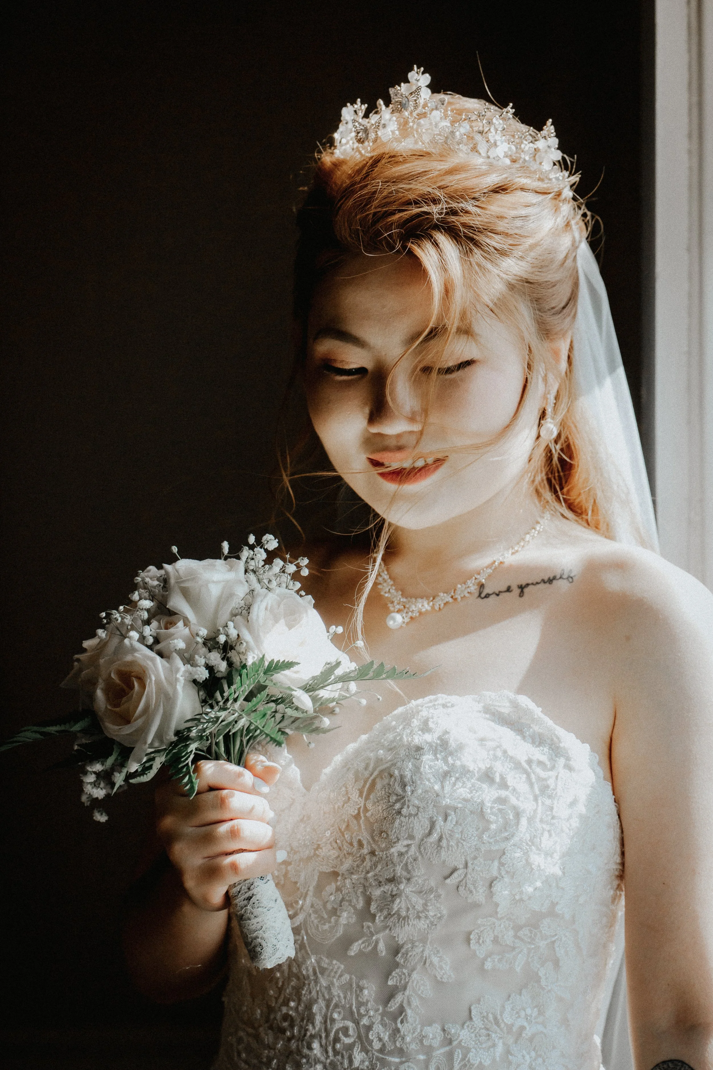 Bride with red hair wearing a tiara, lace wedding gown, and pearl necklace, holding a bouquet of white roses, standing by a window with sunlight.