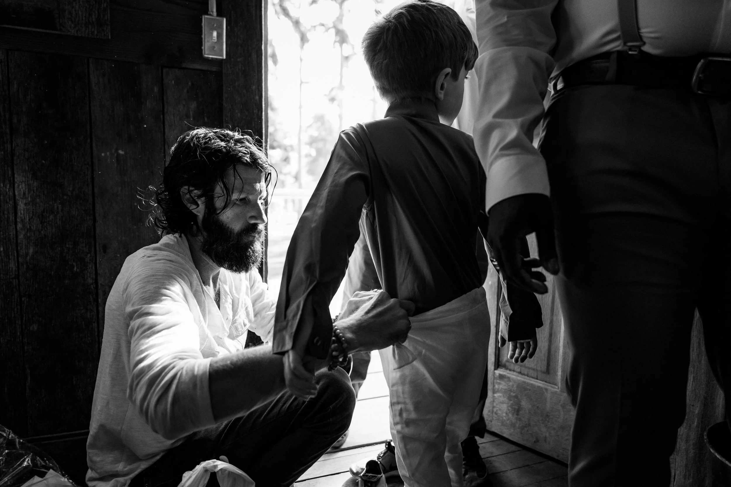 A man with a beard sitting on the floor helping a young boy get dressed, with a third person assisting. The scene takes place indoors by a door with sunlight coming through.