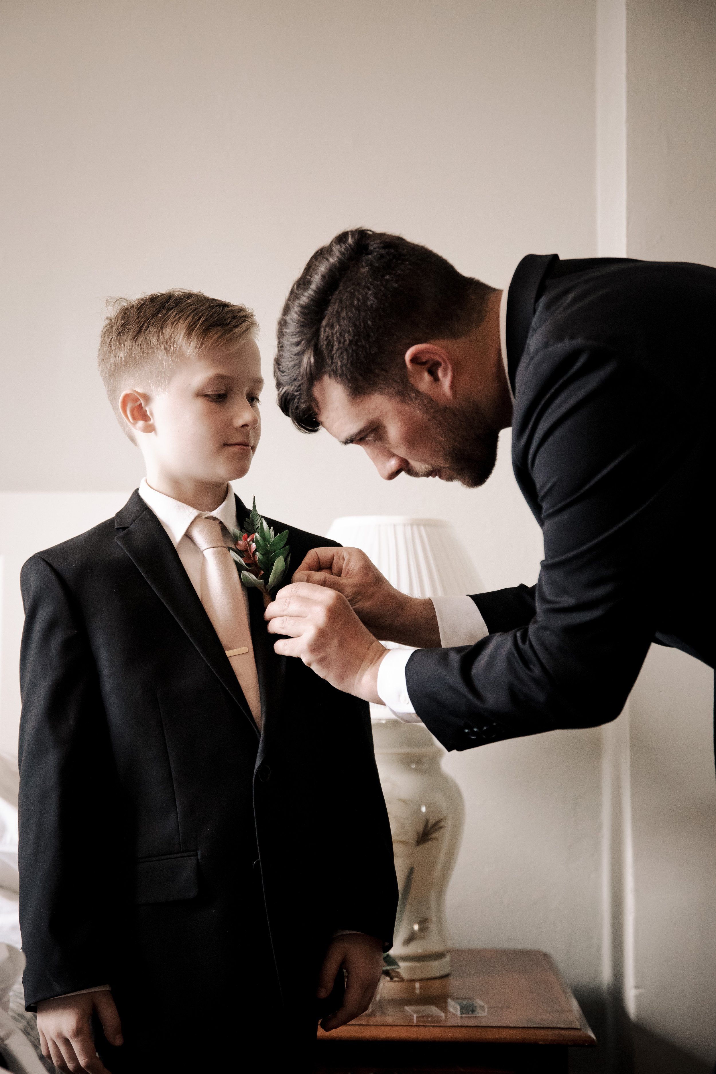 A man in a black suit helping a young boy in a black suit and light pink tie pin a boutonniere on his lapel.