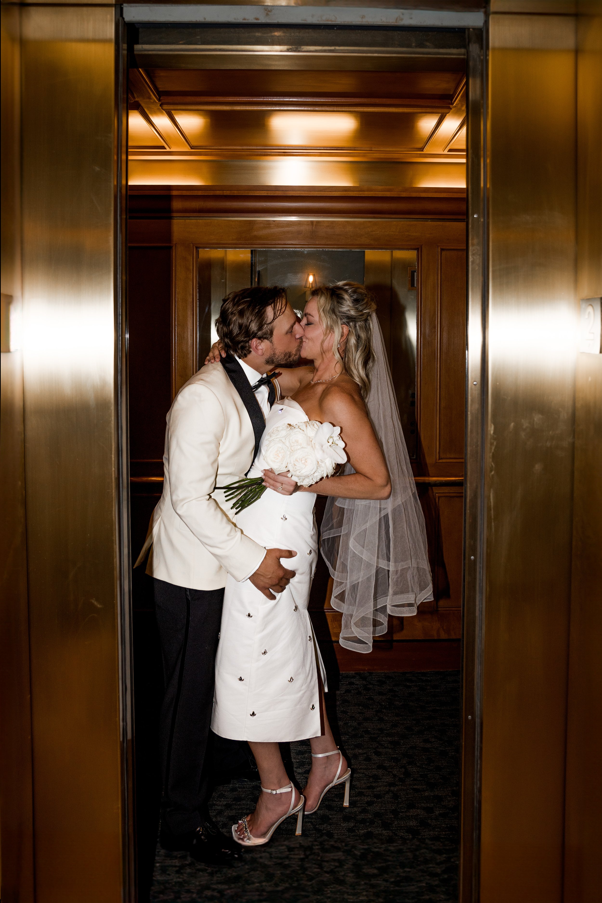 A bride and groom sharing a kiss inside an elevator, with the bride holding a bouquet of white roses.