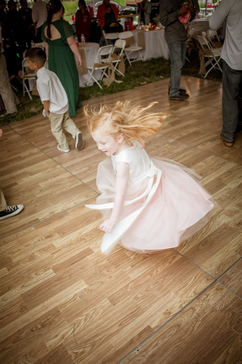 A young girl with red hair spinning on a wooden dance floor at a party or celebration, wearing a light pink dress with a tulle skirt.