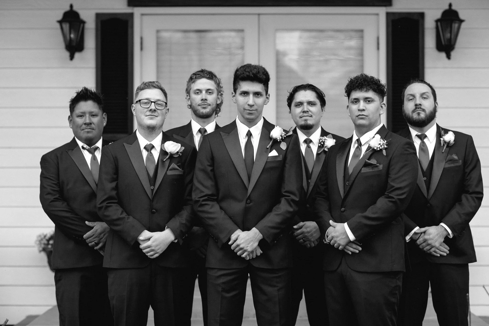 Seven men in tuxedos standing in front of a house, some with boutonnières, posing for a photo.