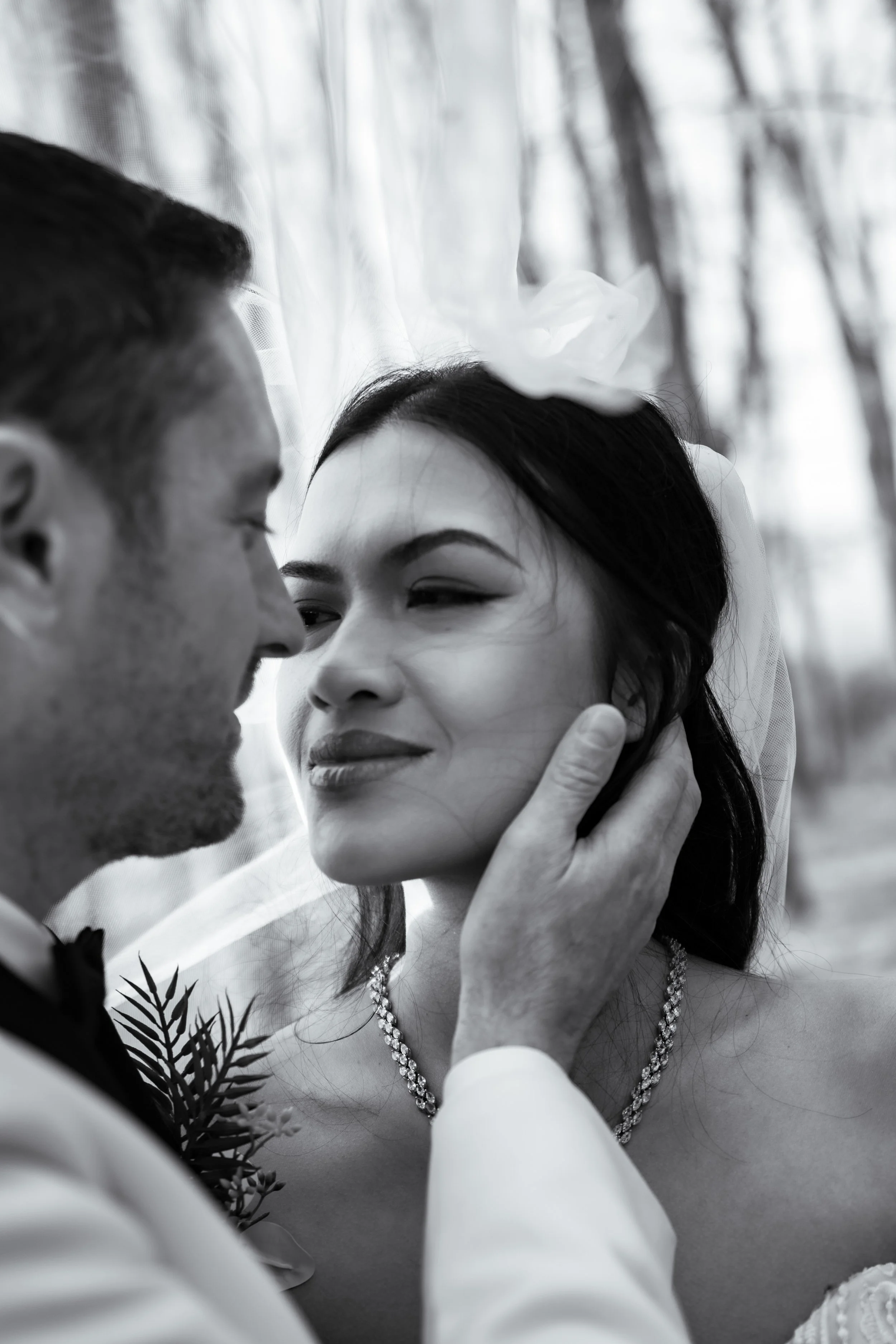A black and white photo of a couple, close-up, on their wedding day. The man holds the woman's face gently as they look at each other, with a veil over the woman's head, and the background appears to be outdoors with trees.
