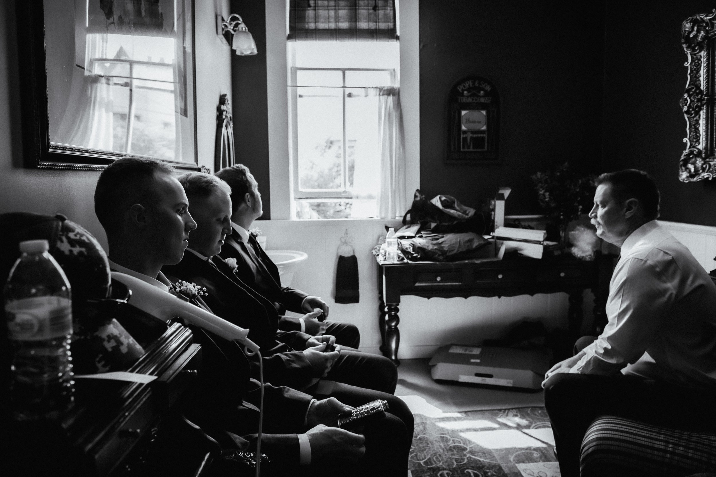 Black and white photo of four young men sitting in a row with hands clasped, facing an older man in a white shirt who is seated on a bench, smoking. The room has sunlight coming through a window and personal belongings on a table in the background.