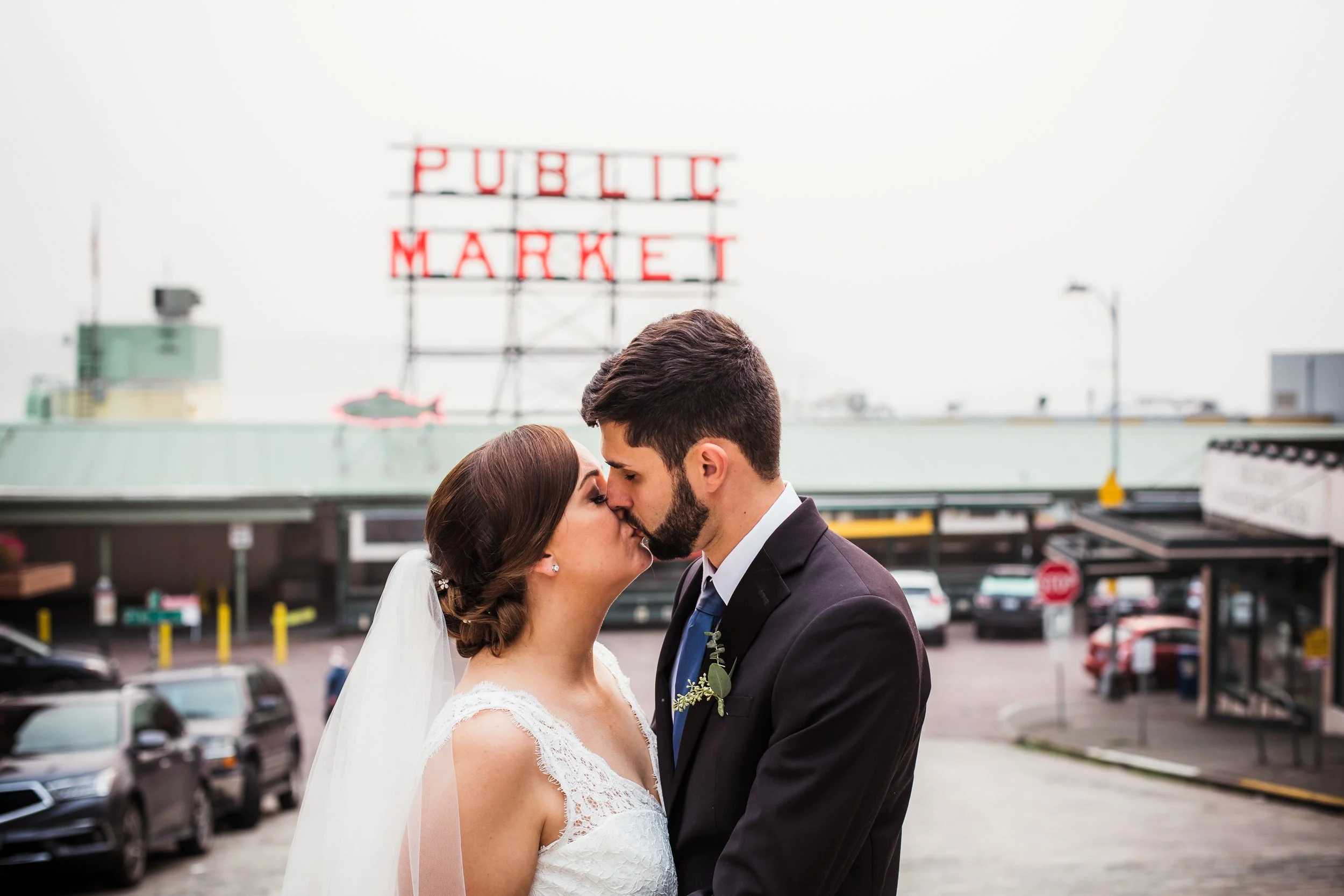 A newlywed couple kissing in front of a public market sign, with cars parked in the background.