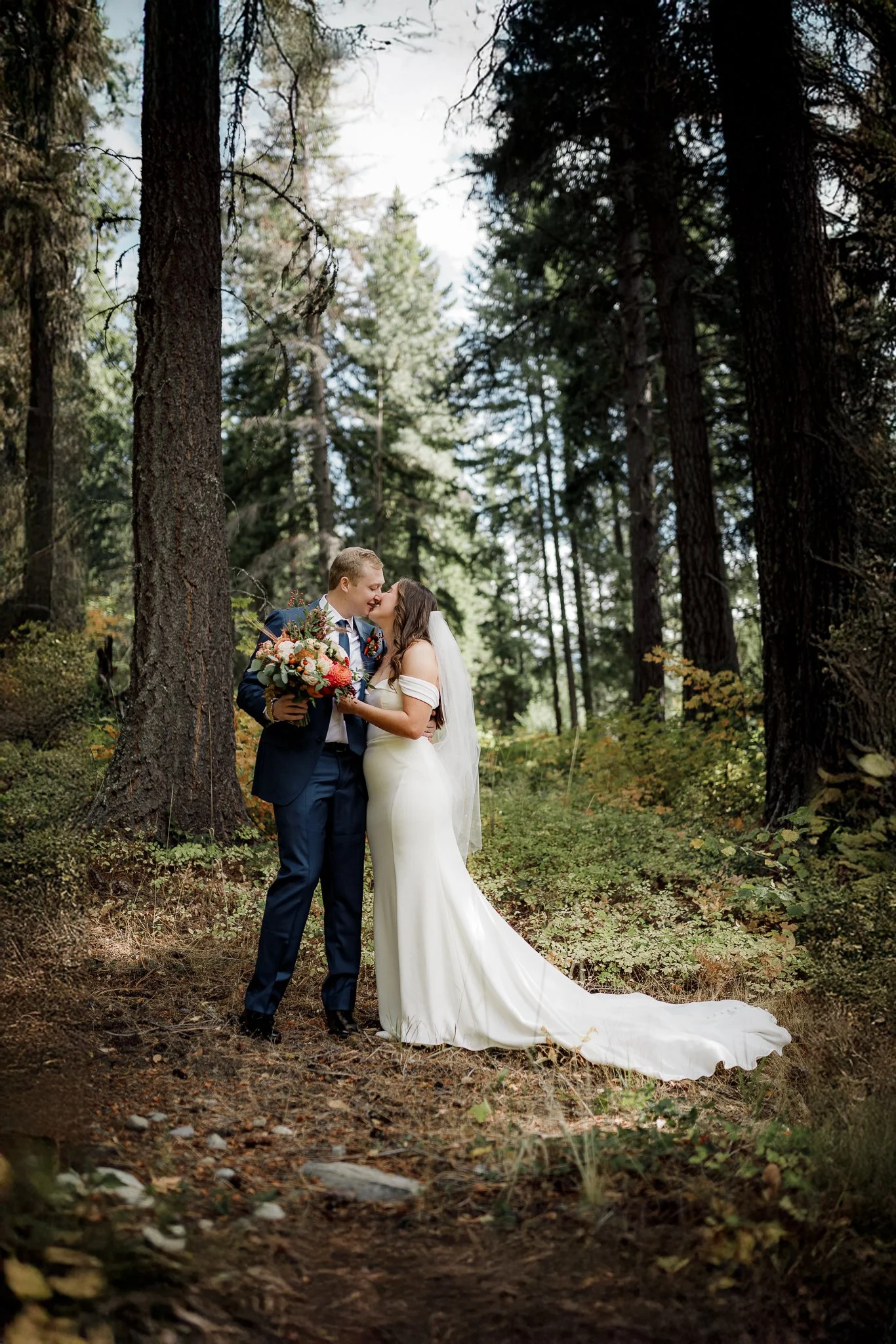 A bride and groom sharing a kiss in a forest during their wedding. The bride is in a white gown and the groom in a dark suit, holding a bouquet of flowers.