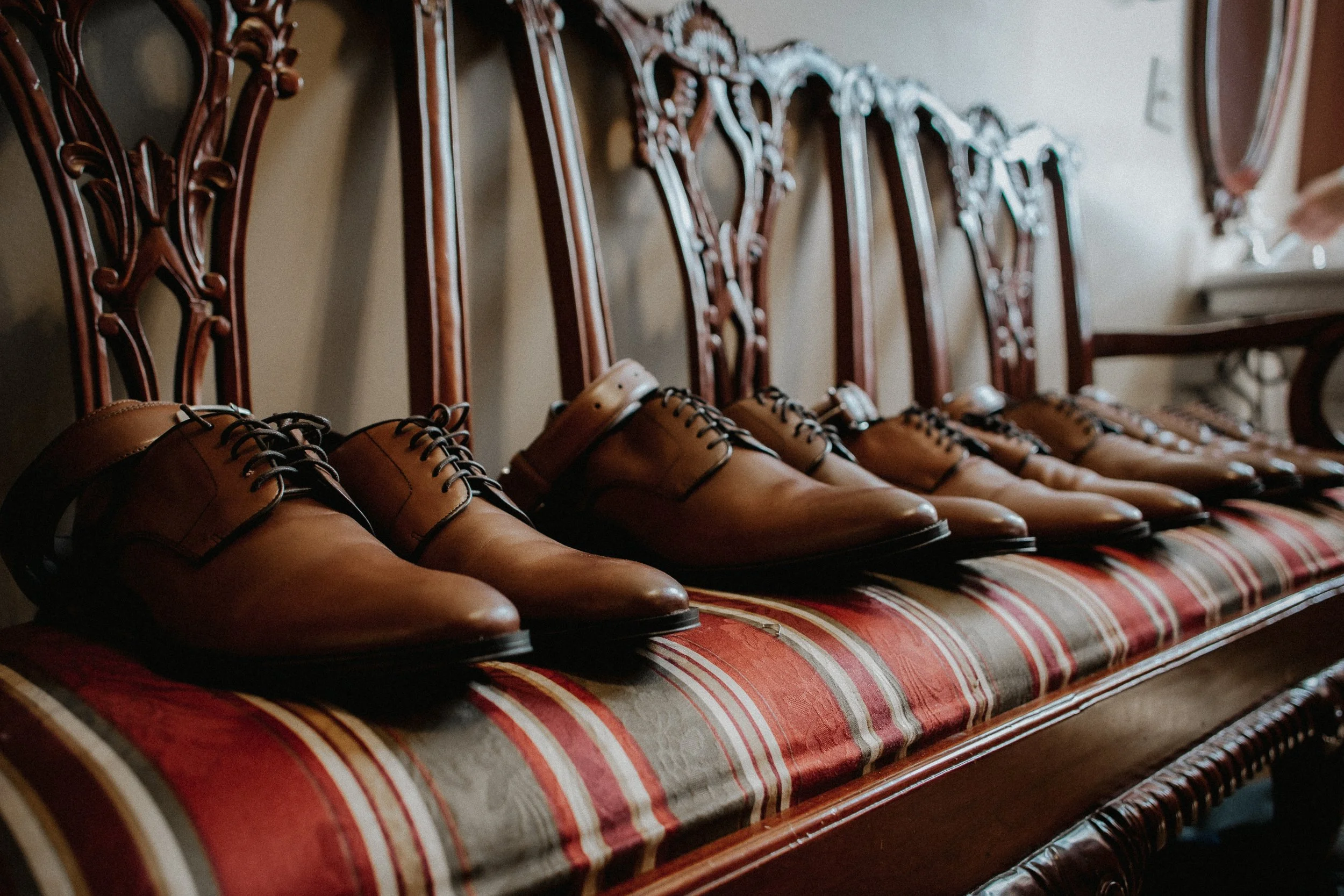 A row of brown leather dress shoes placed on an antique wooden bench with a patterned cushion.