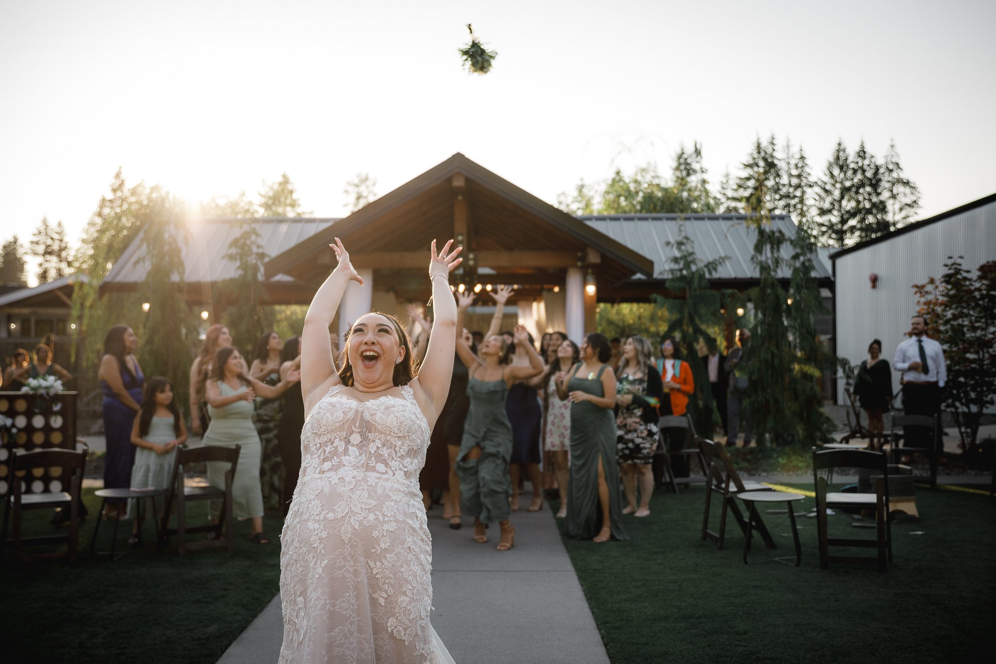 Joyful bride throwing a bouquet at an outdoor wedding reception during sunset, with guests watching and smiling in the background.