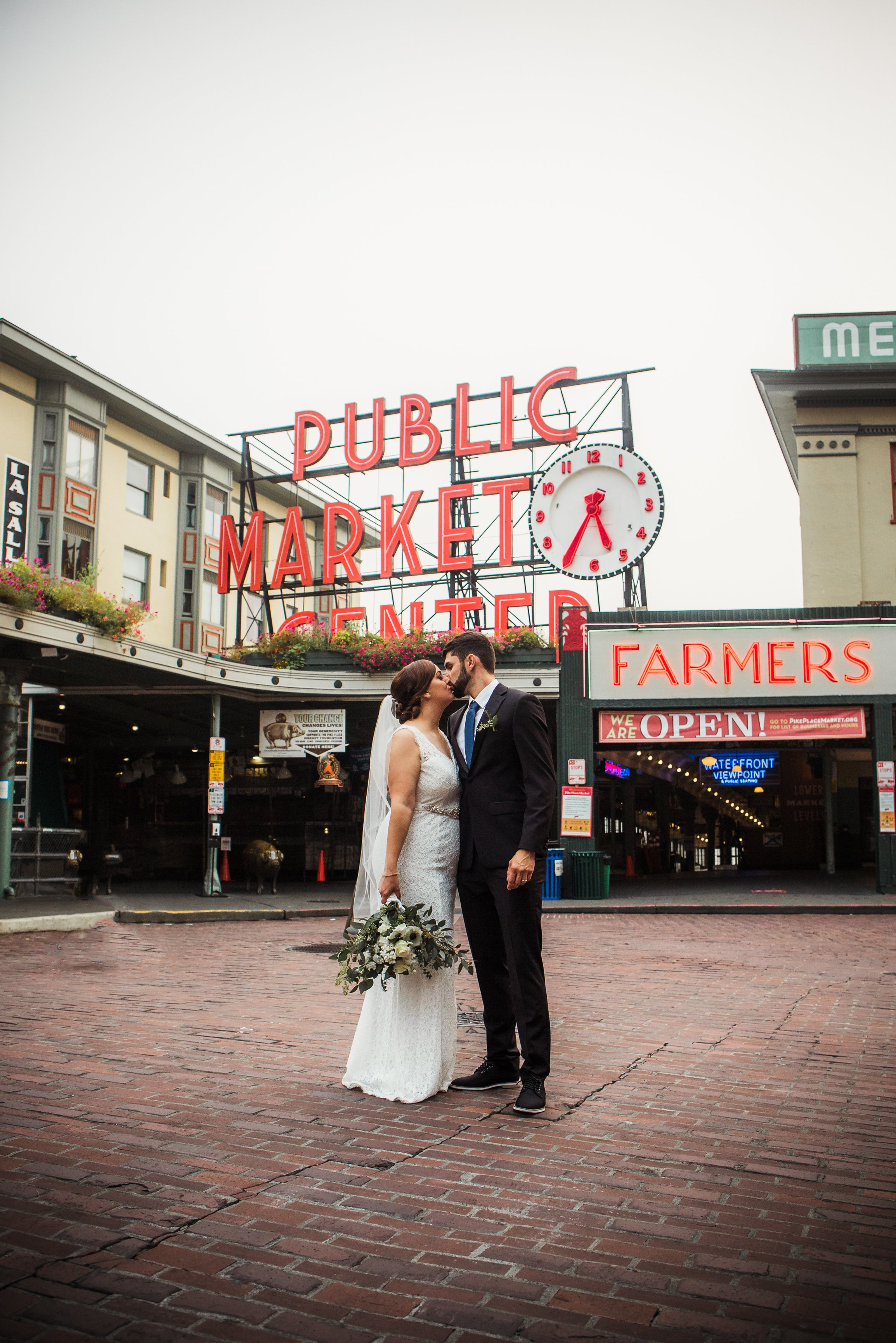 A bride and groom in wedding attire sharing a kiss outside the historic Pike Place Market in Seattle, Washington.