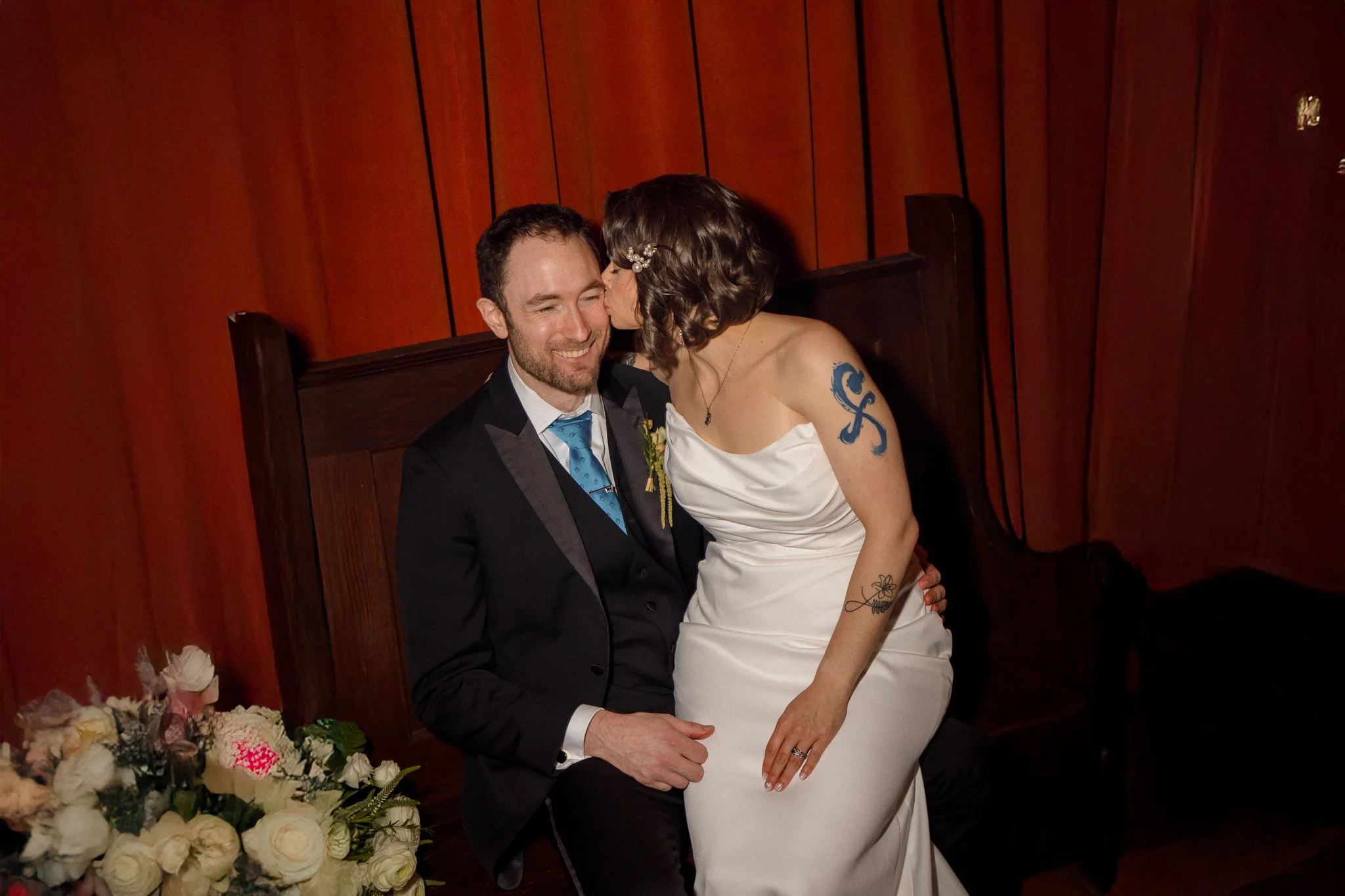 A couple dressed in wedding attire sitting together, with the woman kissing the man on the cheek, in a room with wooden walls and a bouquet of flowers nearby.