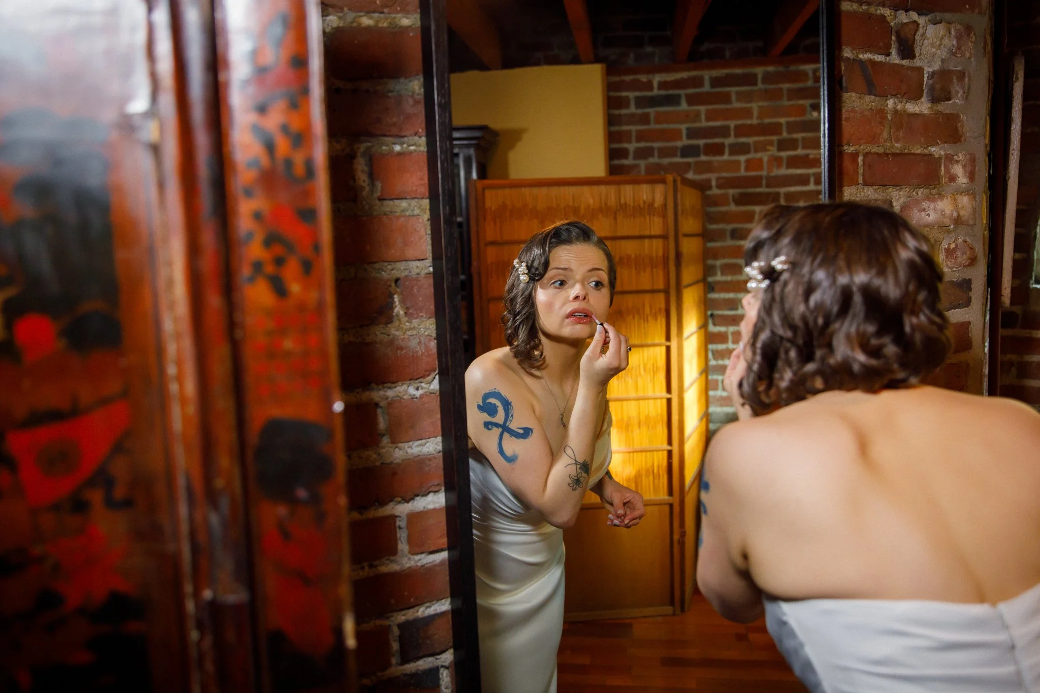 Woman with dark, wavy hair and tattoos on her arm, wearing a strapless dress, applying lipstick while looking into a mirror in a brick-walled room.