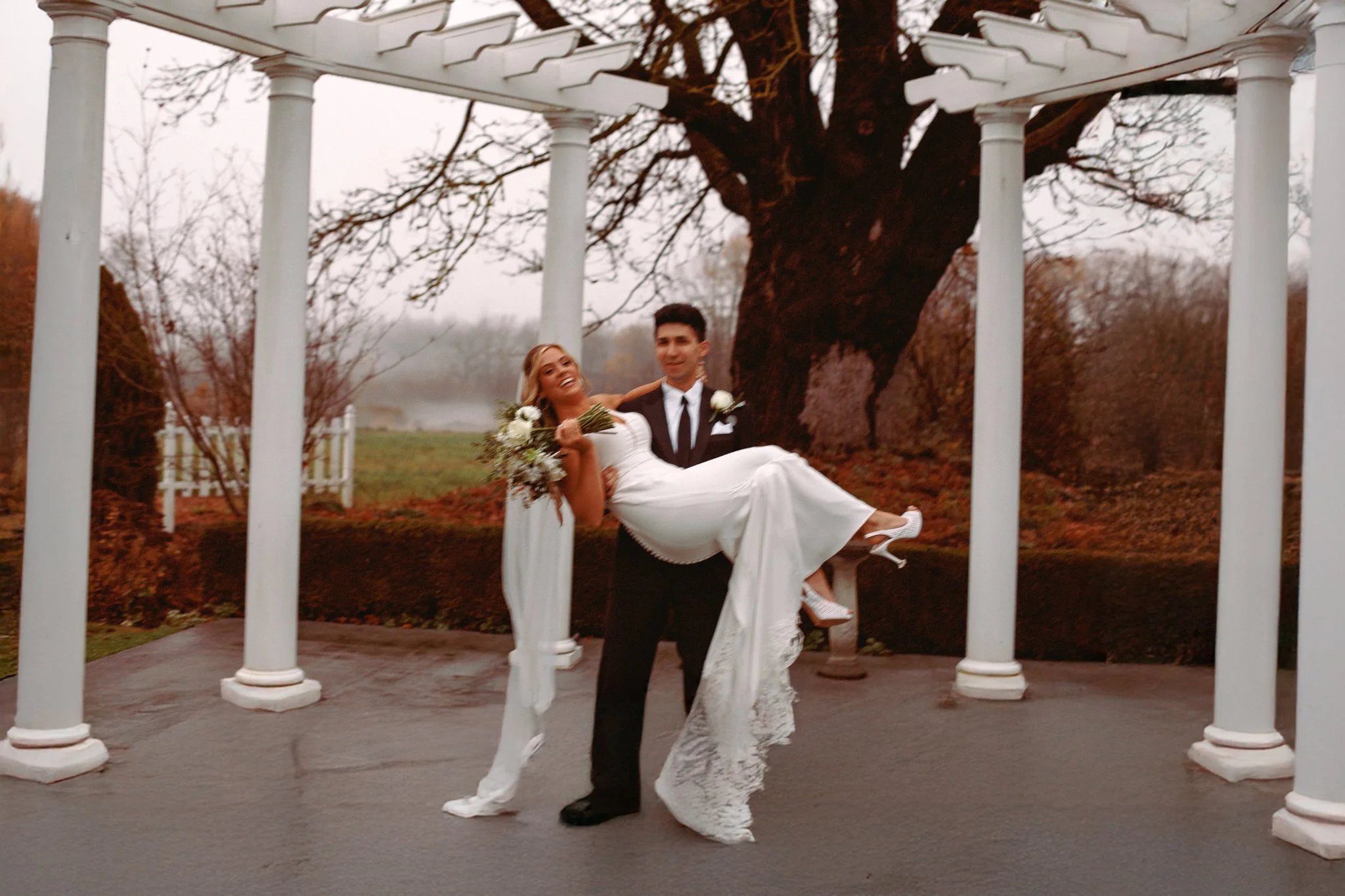 A bride and groom posing under a white pergola outdoors, with the groom holding the bride who is smiling and holding a bouquet of flowers, a large tree in the background, and an overcast sky.