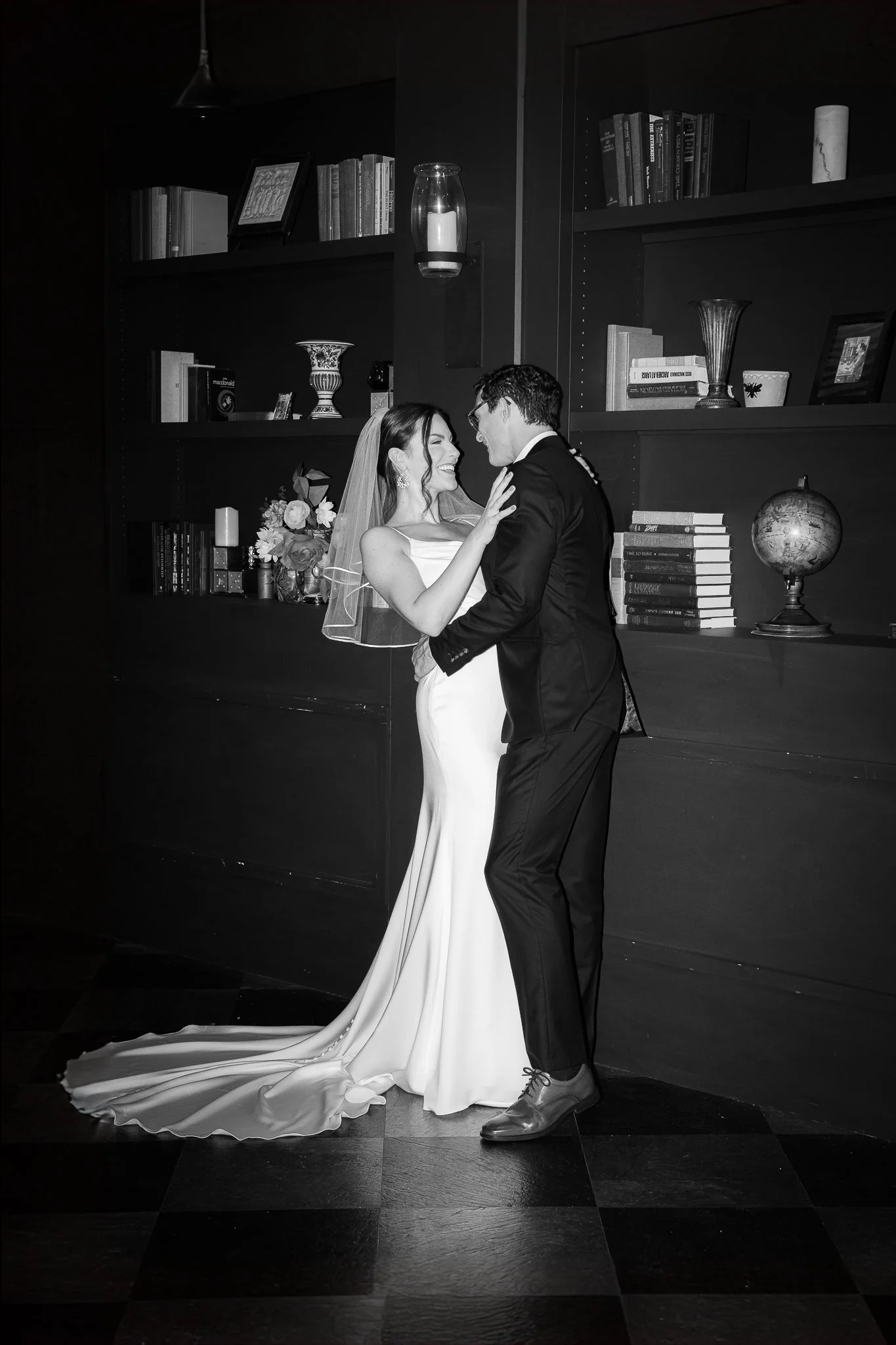 Black and white photo of a bride and groom dancing together in a room with bookshelves and decorative items.