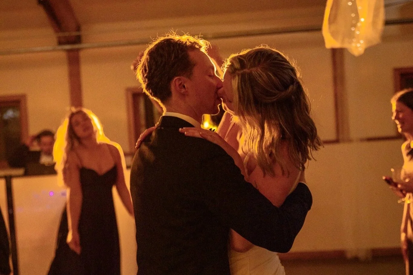 A couple dancing closely at an indoor event, illuminated by warm lighting, with other people in the background.