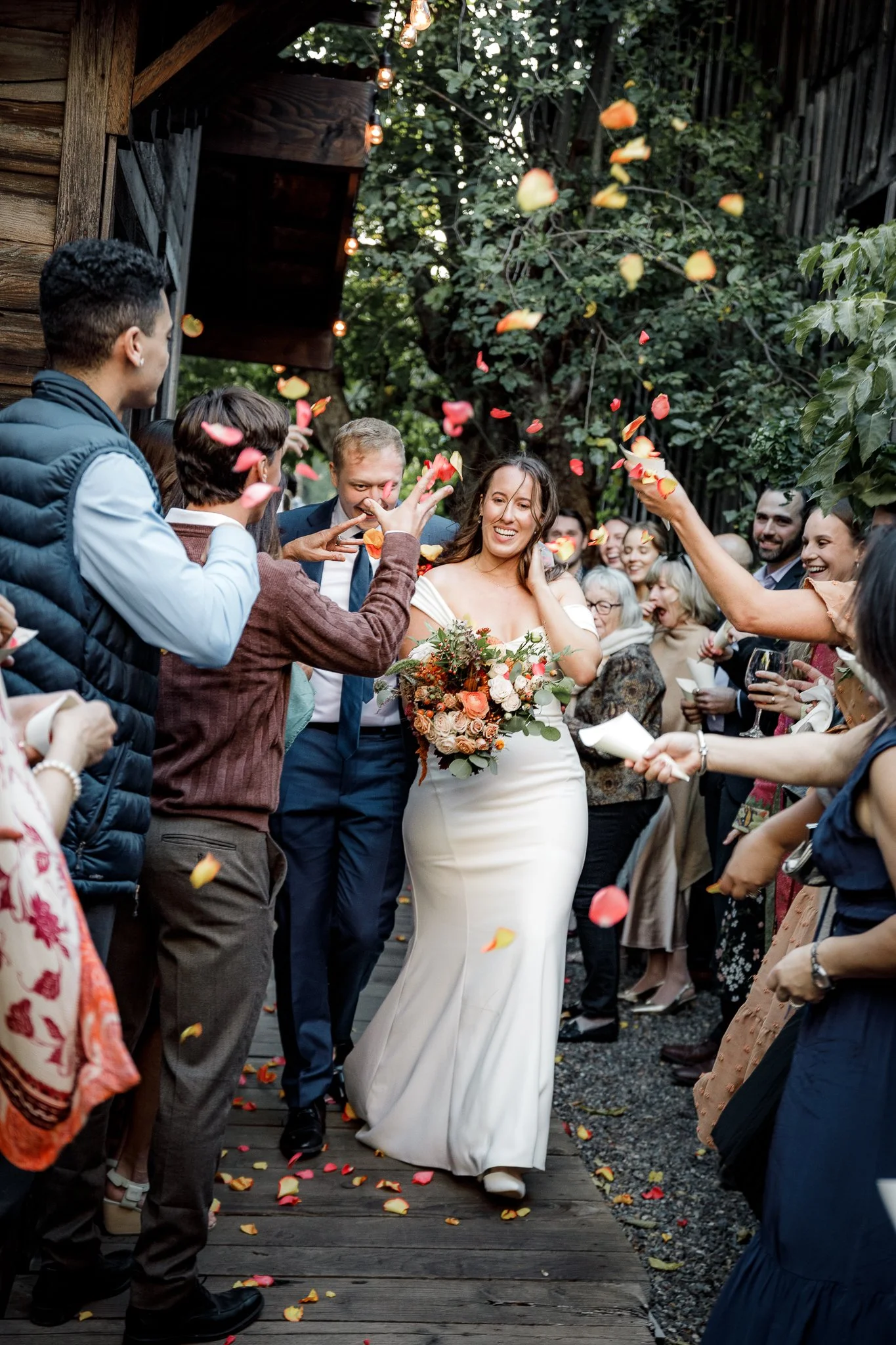 Wedding celebration with guests throwing petals around a bride dressed in white, holding a bouquet, smiling as she walks through an outdoor archway, surrounded by trees and wooden structures.