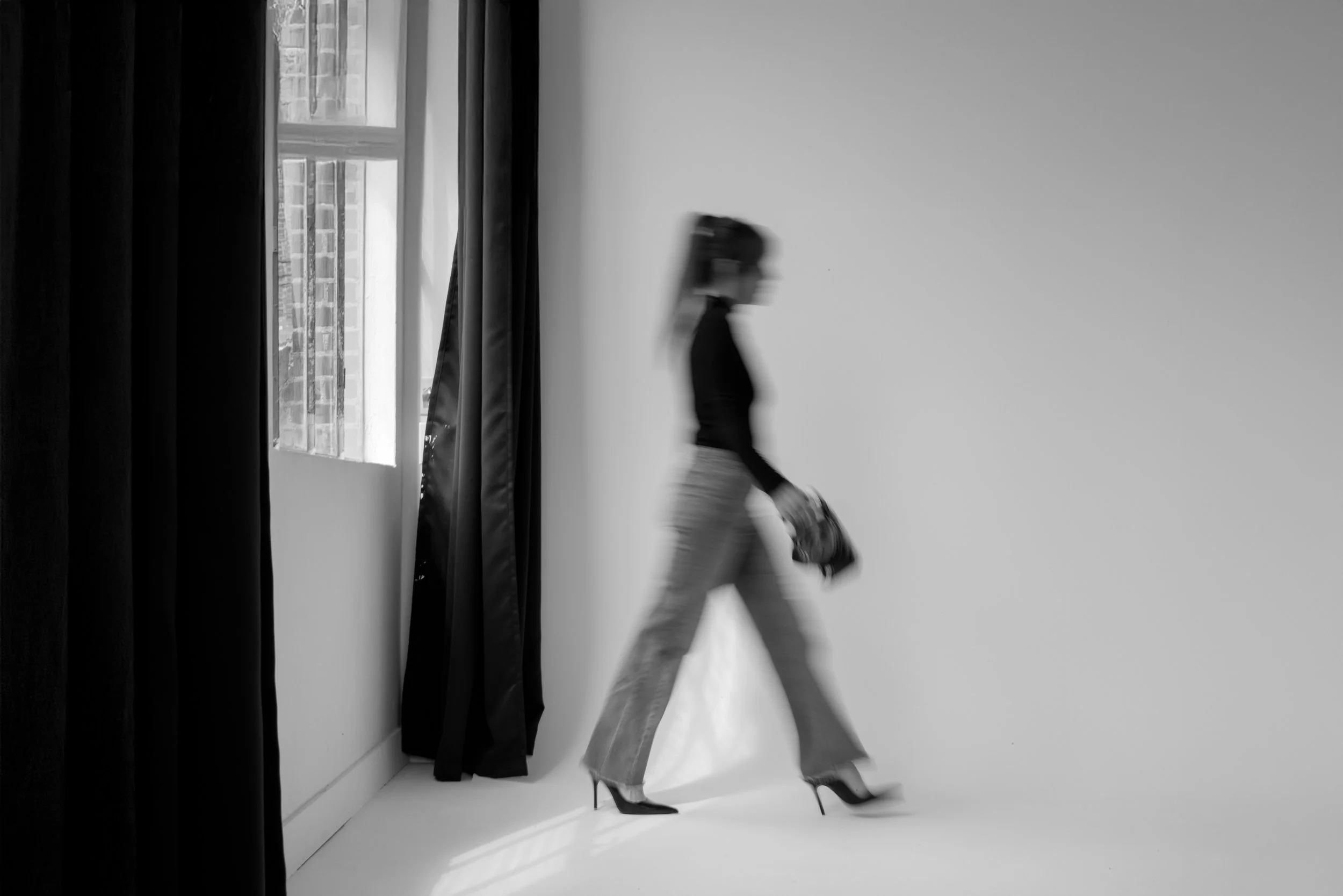 A woman walking past a window with black curtains, wearing high-heeled shoes, jeans, and a long-sleeved top, with her hair tied back.