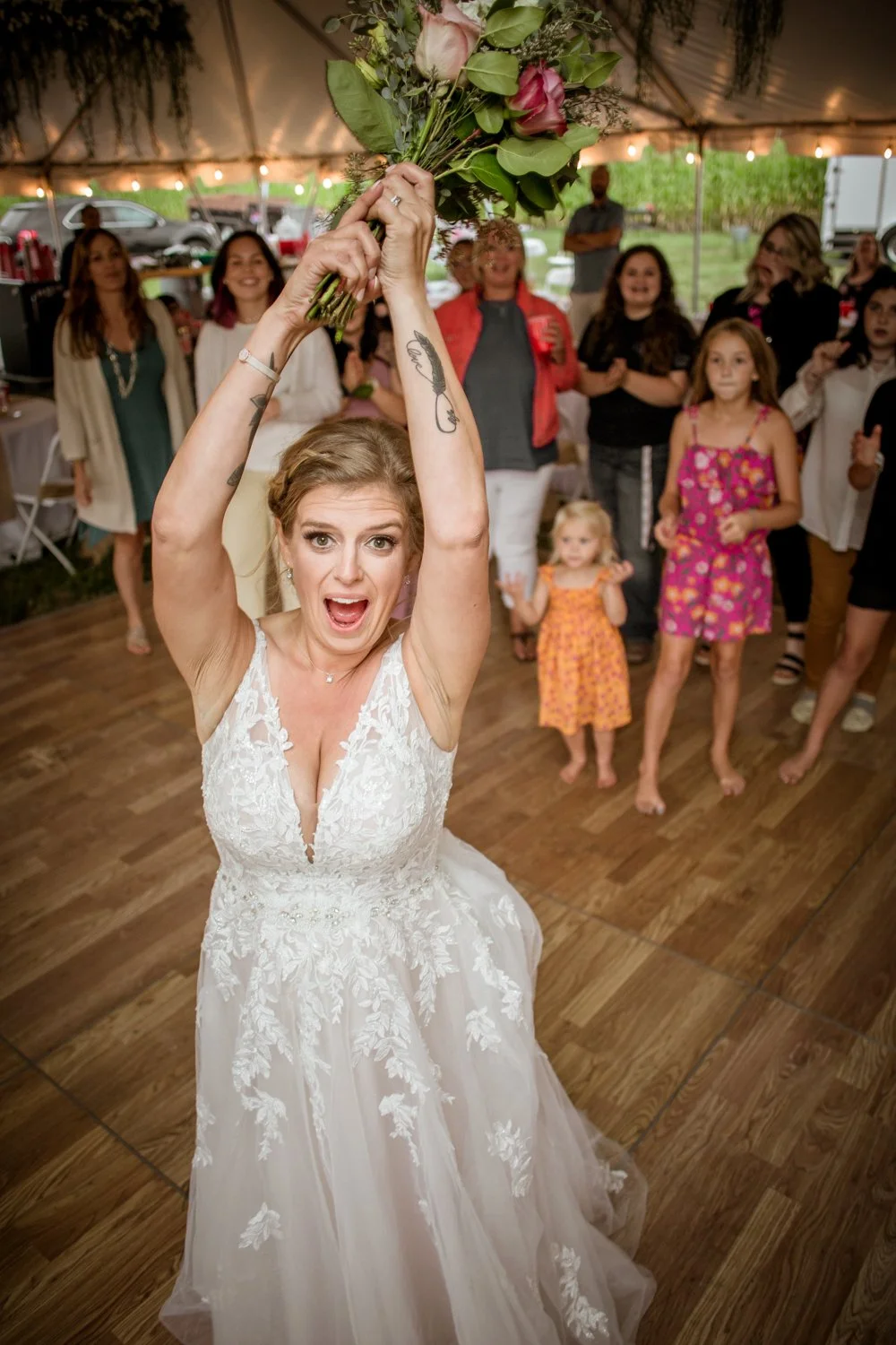 Bride swinging a bouquet during a wedding reception in a tent with guests watching and smiling.