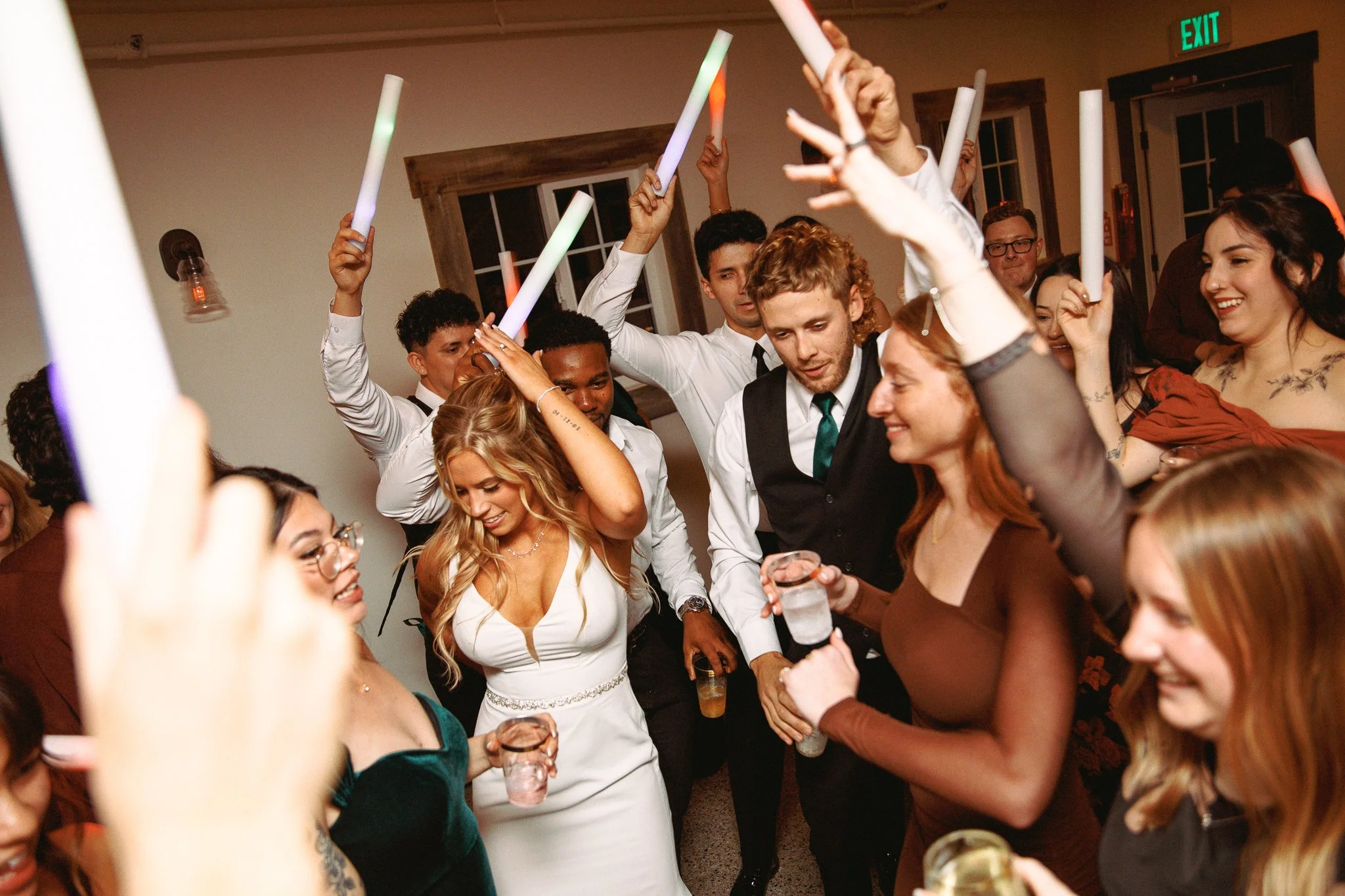 Group of people celebrating, some holding glow sticks, dancing happy at a party or wedding reception.