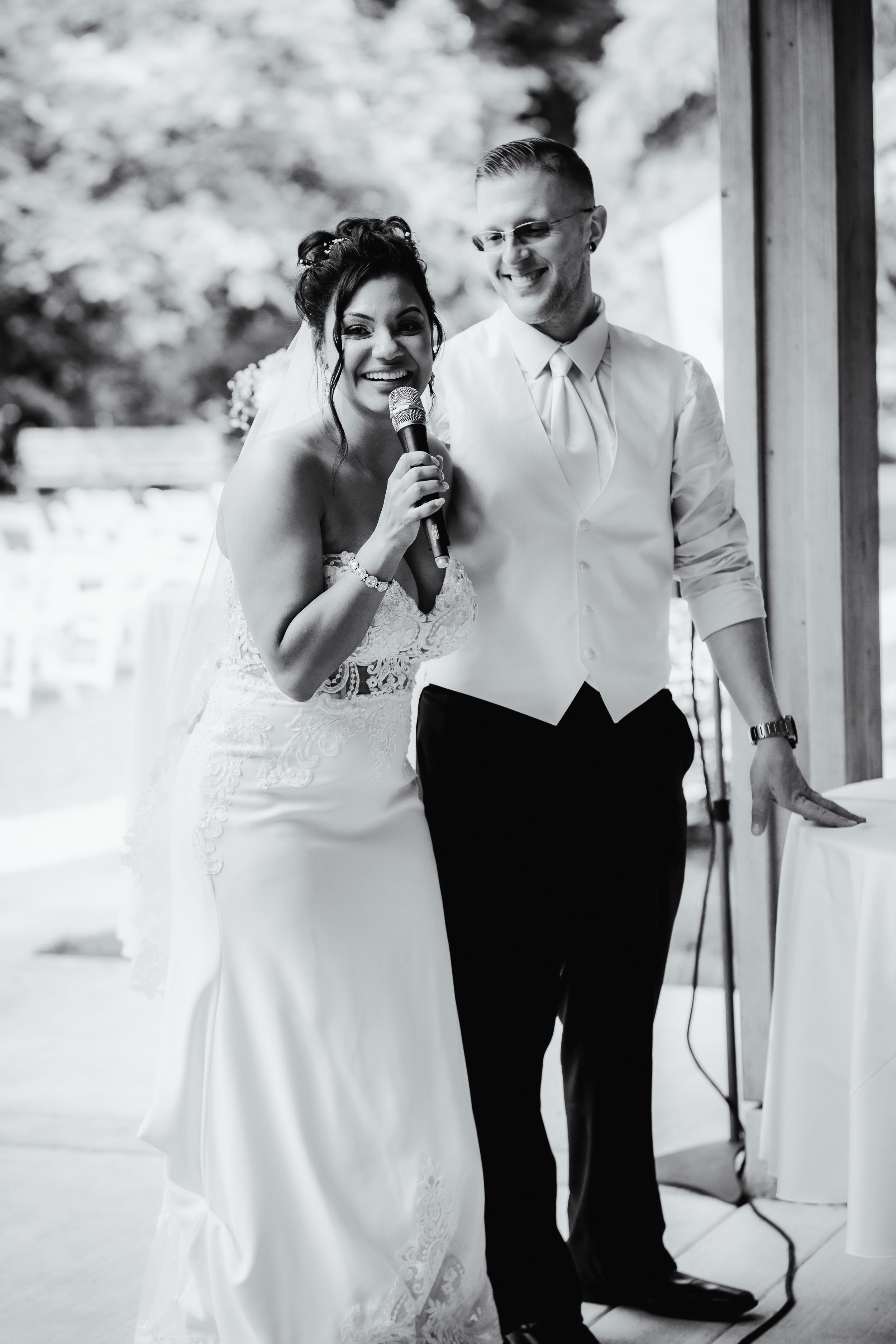A bride and groom smiling at their wedding reception, with the bride holding a microphone. The bride is wearing a lace wedding dress and the groom is dressed in a white shirt with a vest, standing close together indoors.