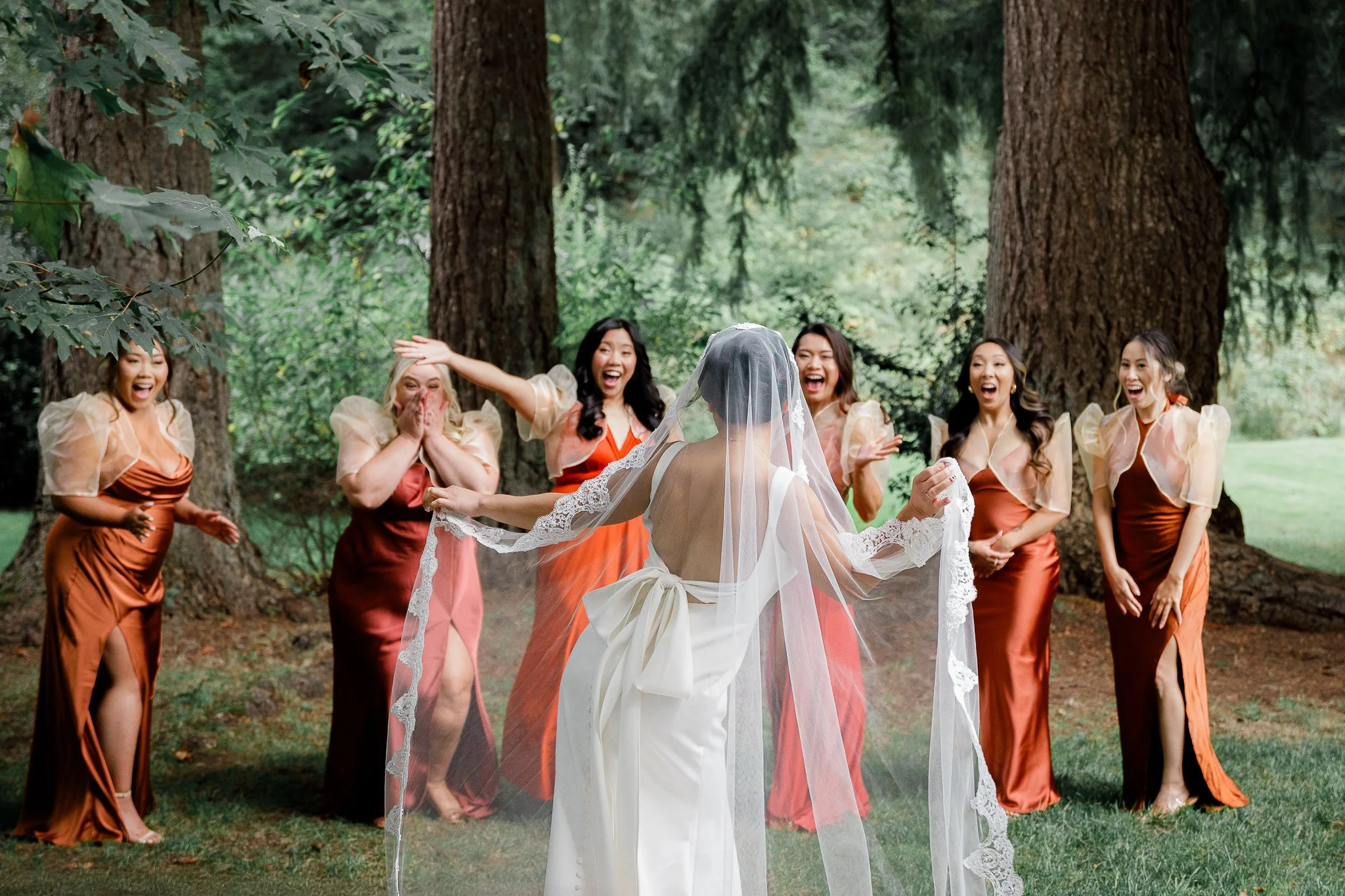 Bride in a white wedding dress with a veil holding hands with six bridesmaids in burnt orange dresses, all laughing and joyful in a wooded outdoor setting.