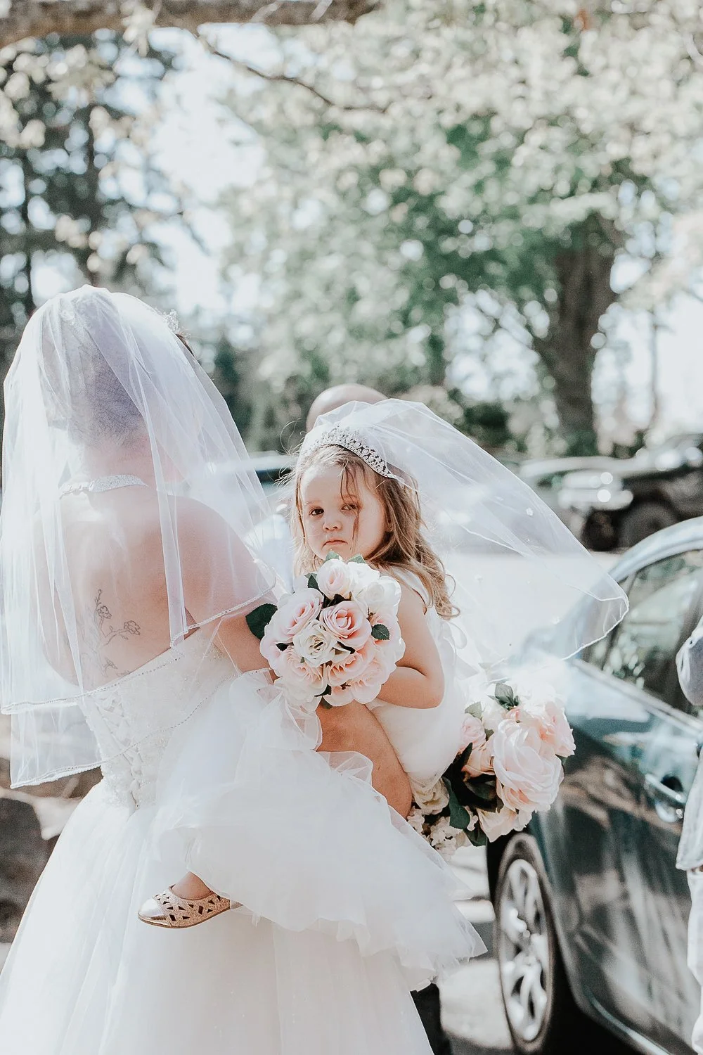 A bride holding a young girl wearing a white dress and veil, both holding bouquets of pink and white flowers, outdoors with parked cars and trees in the background.