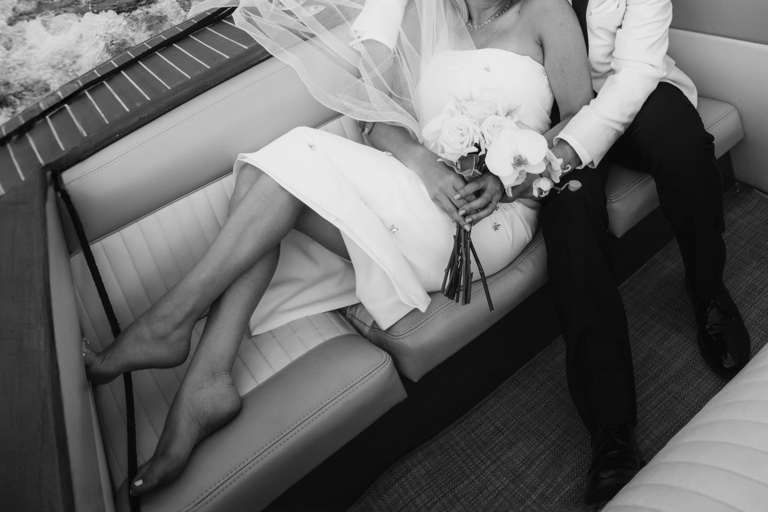 A bride and groom sitting close together on a boat, with their hands intertwined and the bride holding a bouquet. The bride is in a strapless dress, and the groom is wearing a white blazer and black pants.