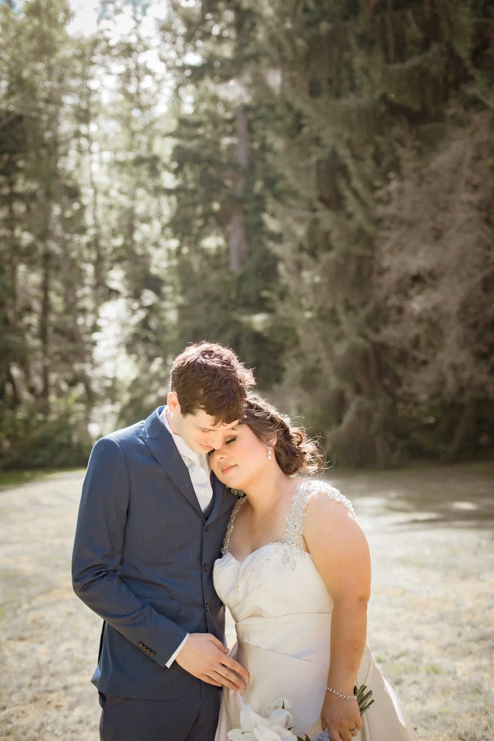 A bride and groom share a tender moment outdoors, with the bride holding a bouquet and the groom wearing a navy suit, surrounded by trees and natural light.