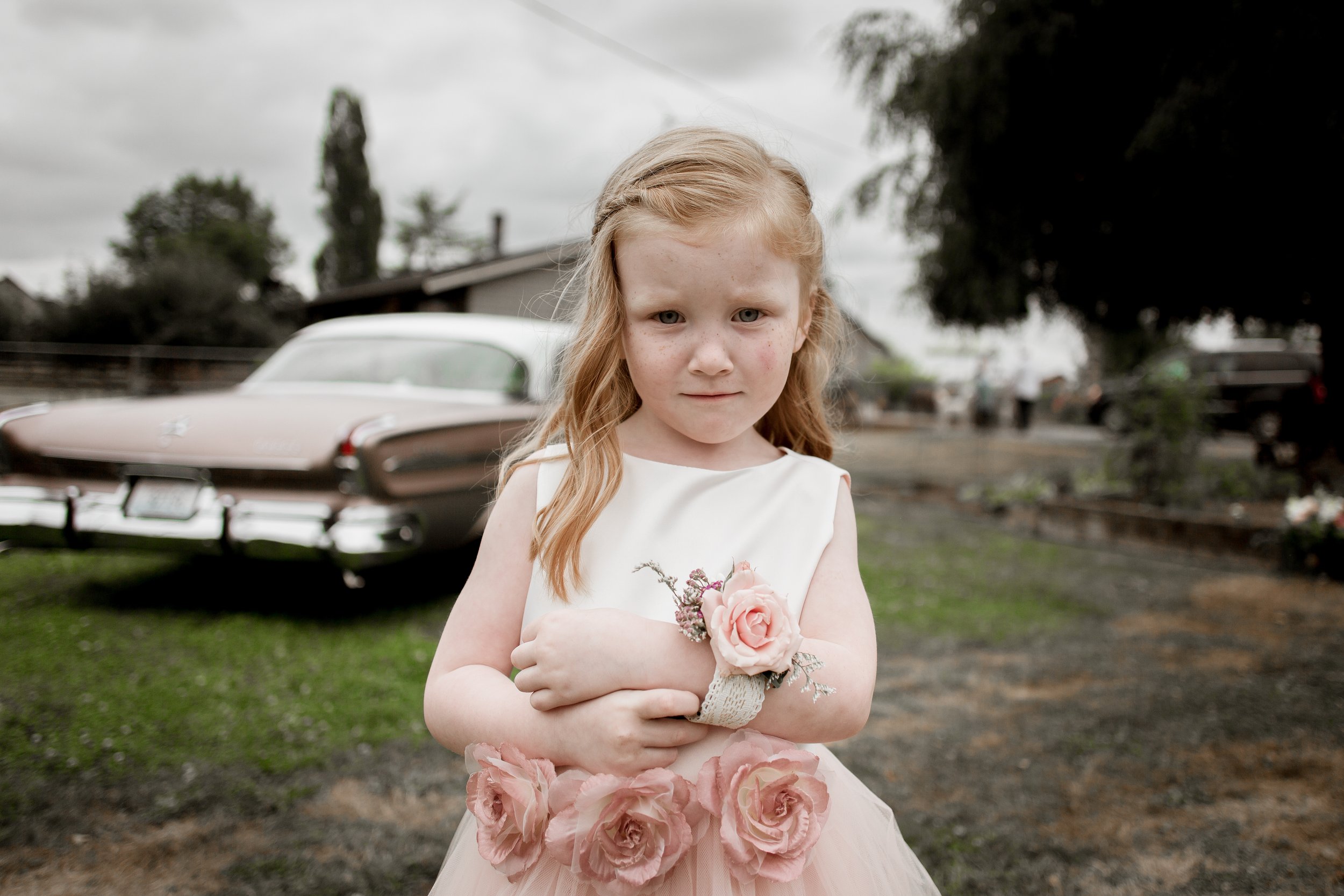 A young girl with red hair standing outdoors on a cloudy day, holding a pink flower corsage in front of her with a neutral expression. She is wearing a cream-colored dress with pink roses on the skirt and wrist corsage, and is standing in front of a 