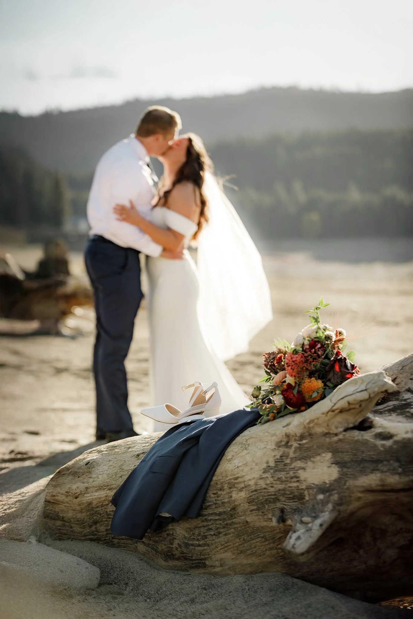 A bride and groom kissing on a beach, with a wedding bouquet, navy blue suit, and white high heels placed on a large piece of driftwood in the foreground.