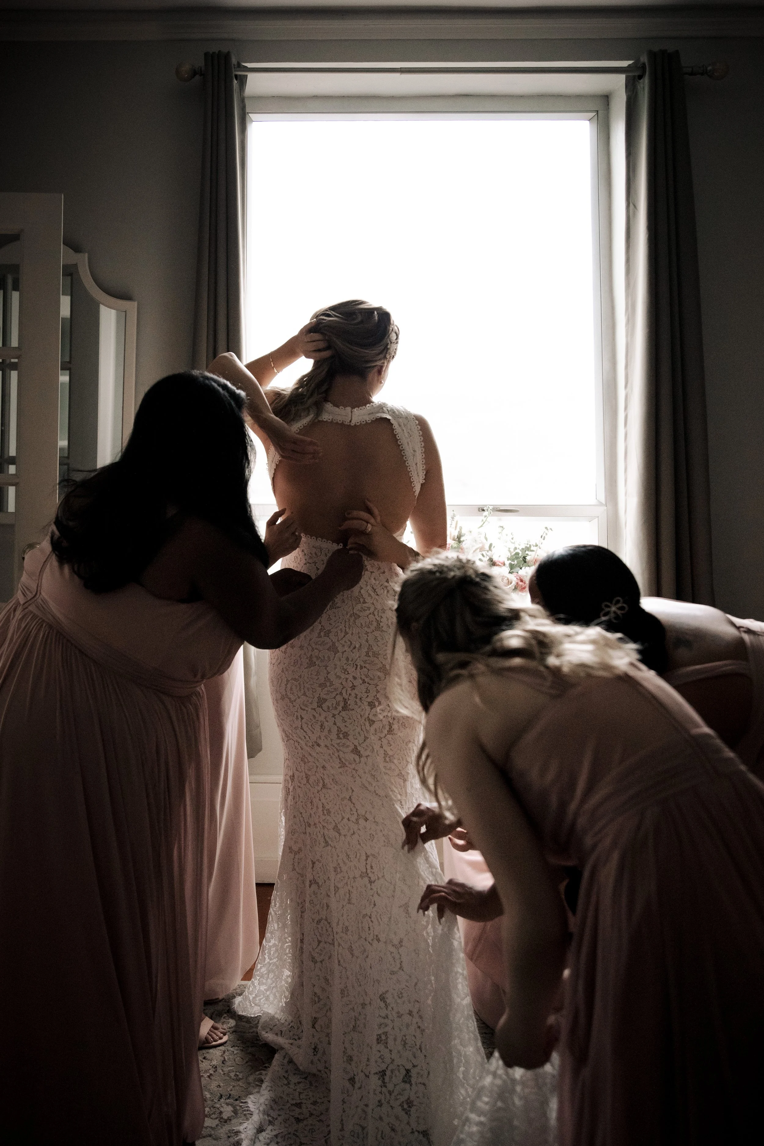 A bride in a lace wedding dress is getting help from bridesmaids as they prepare inside a room near a window with curtains.