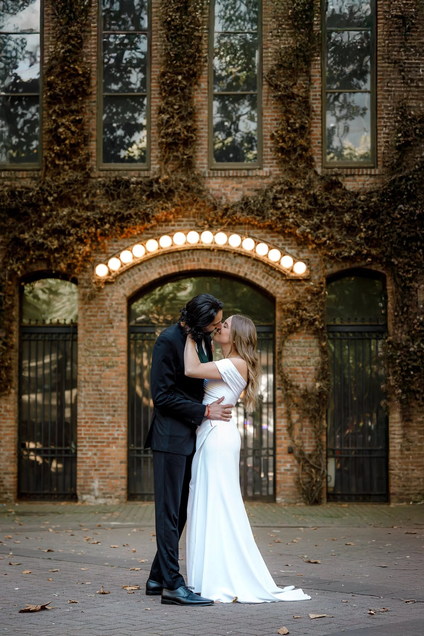 A couple in wedding attire sharing a kiss outdoors in front of a brick building with ivy and large windows