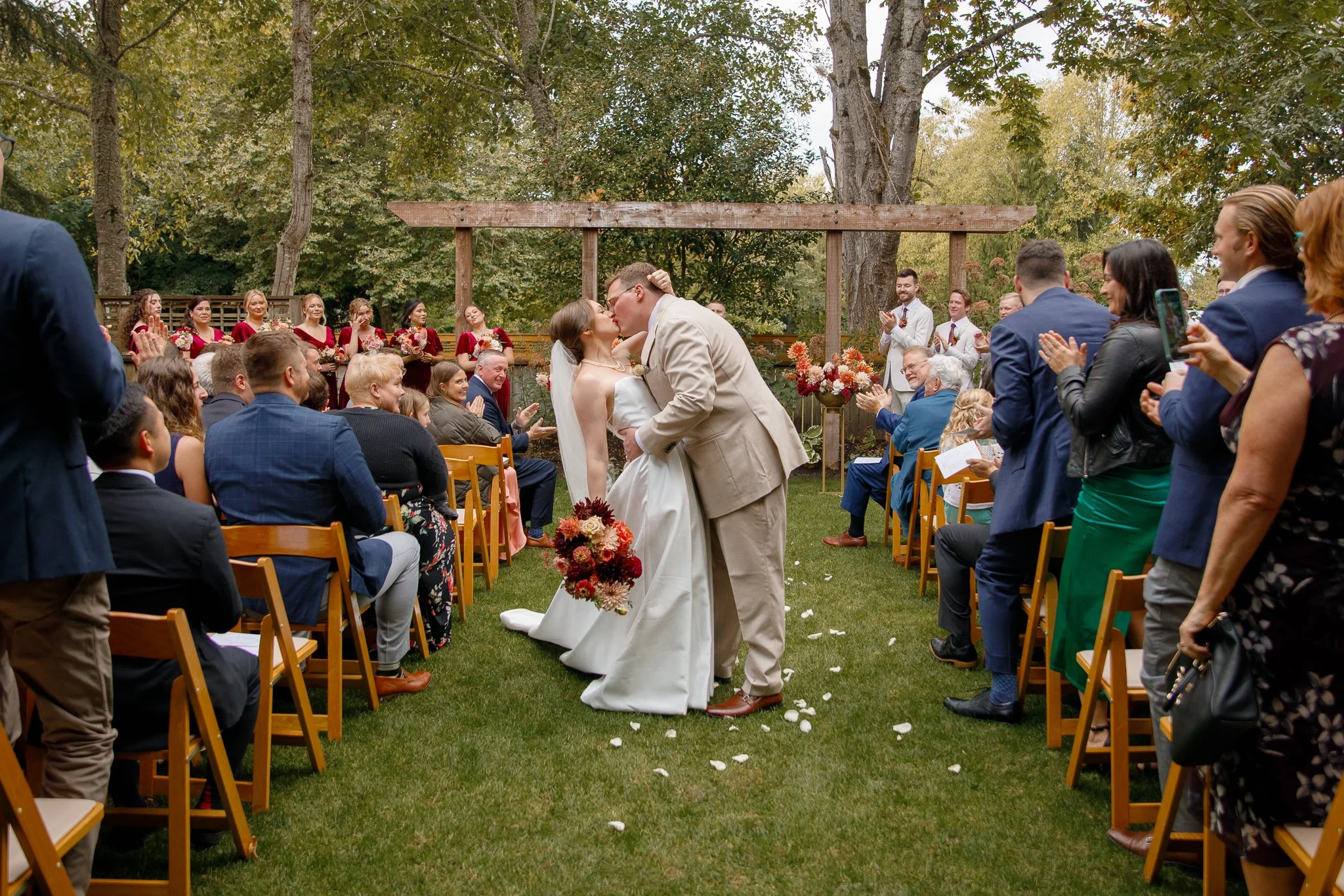 A couple shares a kiss during their outdoor wedding ceremony, surrounded by seated guests applauding and capturing the moment. Bridesmaids dressed in red stand in the background, with a wooden arch and floral arrangements behind them. The setting is a lush green garden with trees.