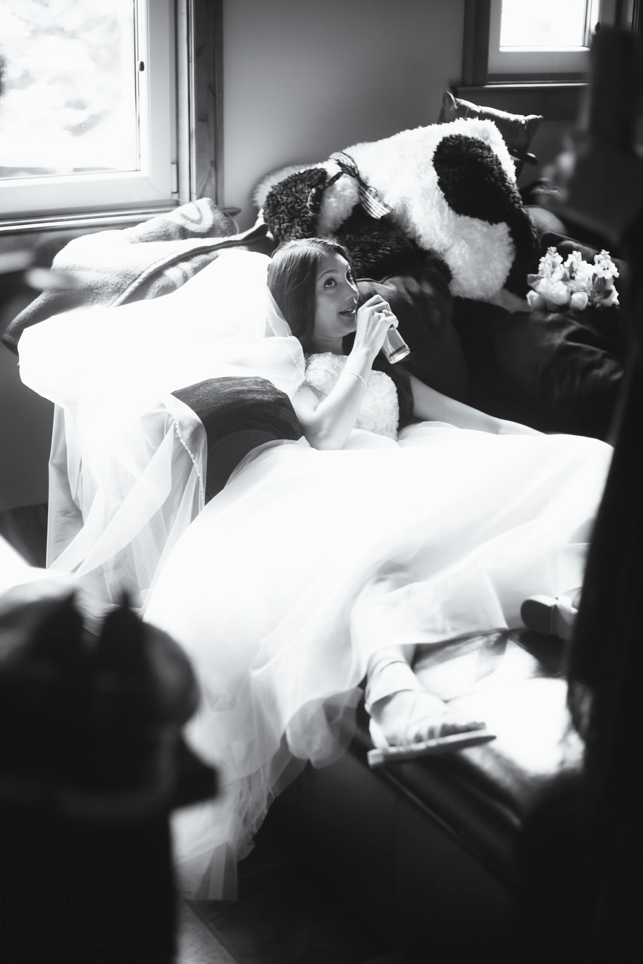 Black and white photo of a bride lying on a couch, holding a drink, with her head on a pillow. There are blankets and pillows on the couch, and a window in the background.