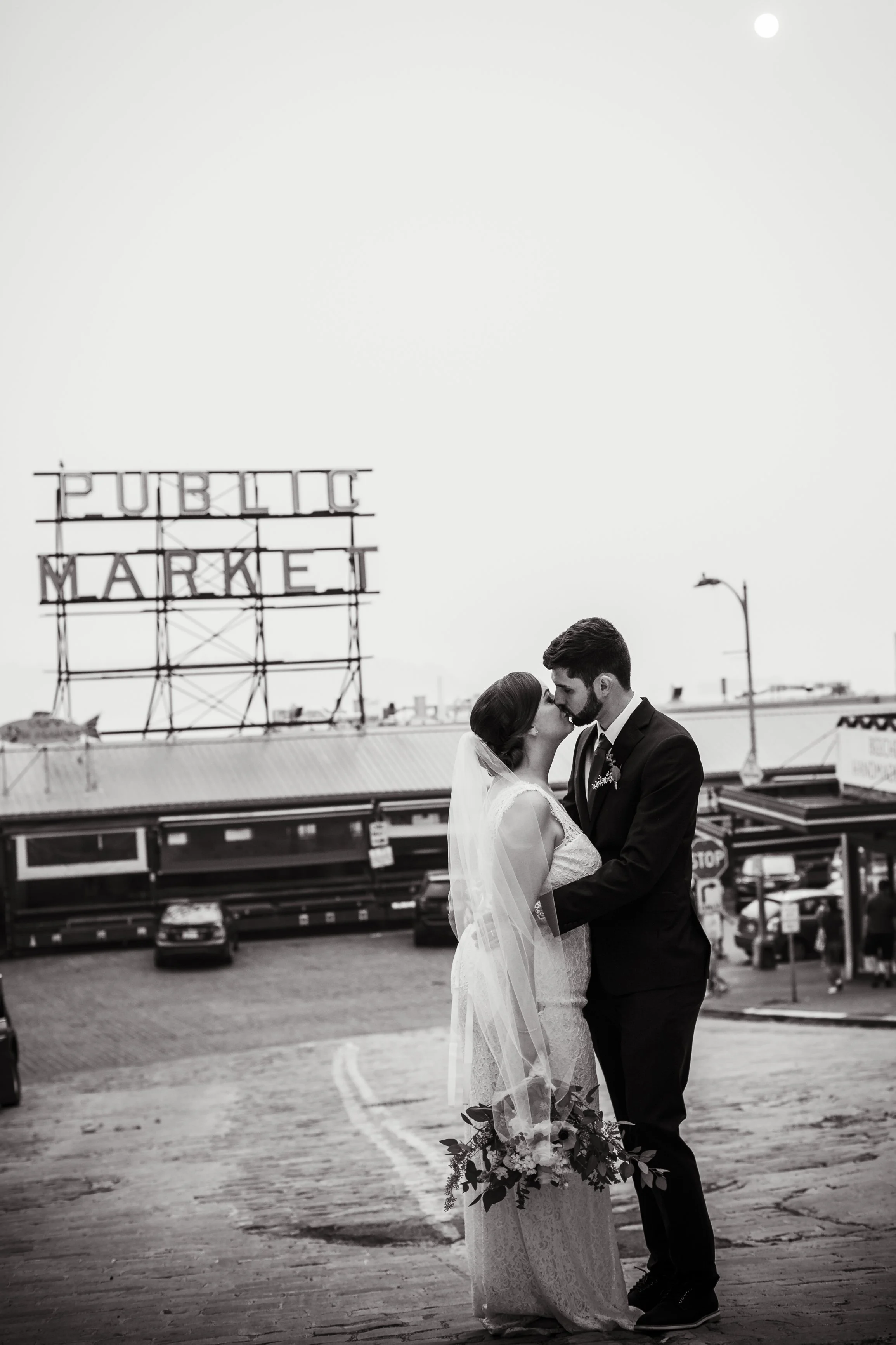 A black and white photo of a bride and groom kissing in a parking lot with a sign reading 'PUBLIC MARKET' in the background.