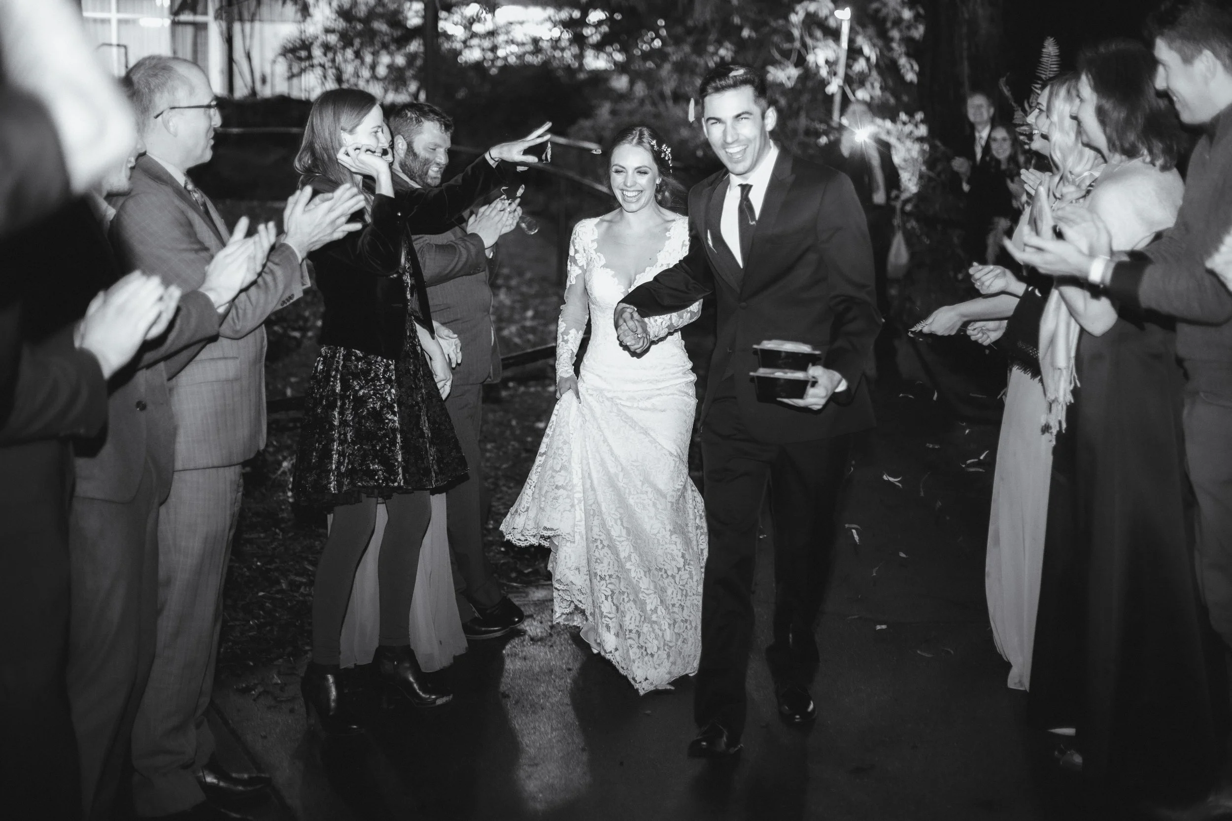 A wedding celebration with a bride and groom walking through a tunnel of friends and family who are clapping and smiling outdoors at night.